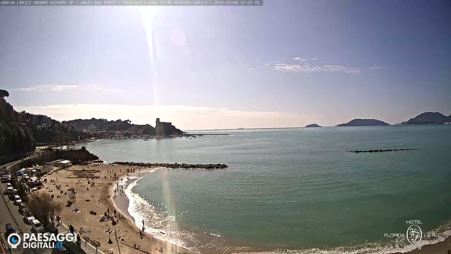 A sunny day at a sandy beach with calm, teal water, overlooking a town and distant hills.