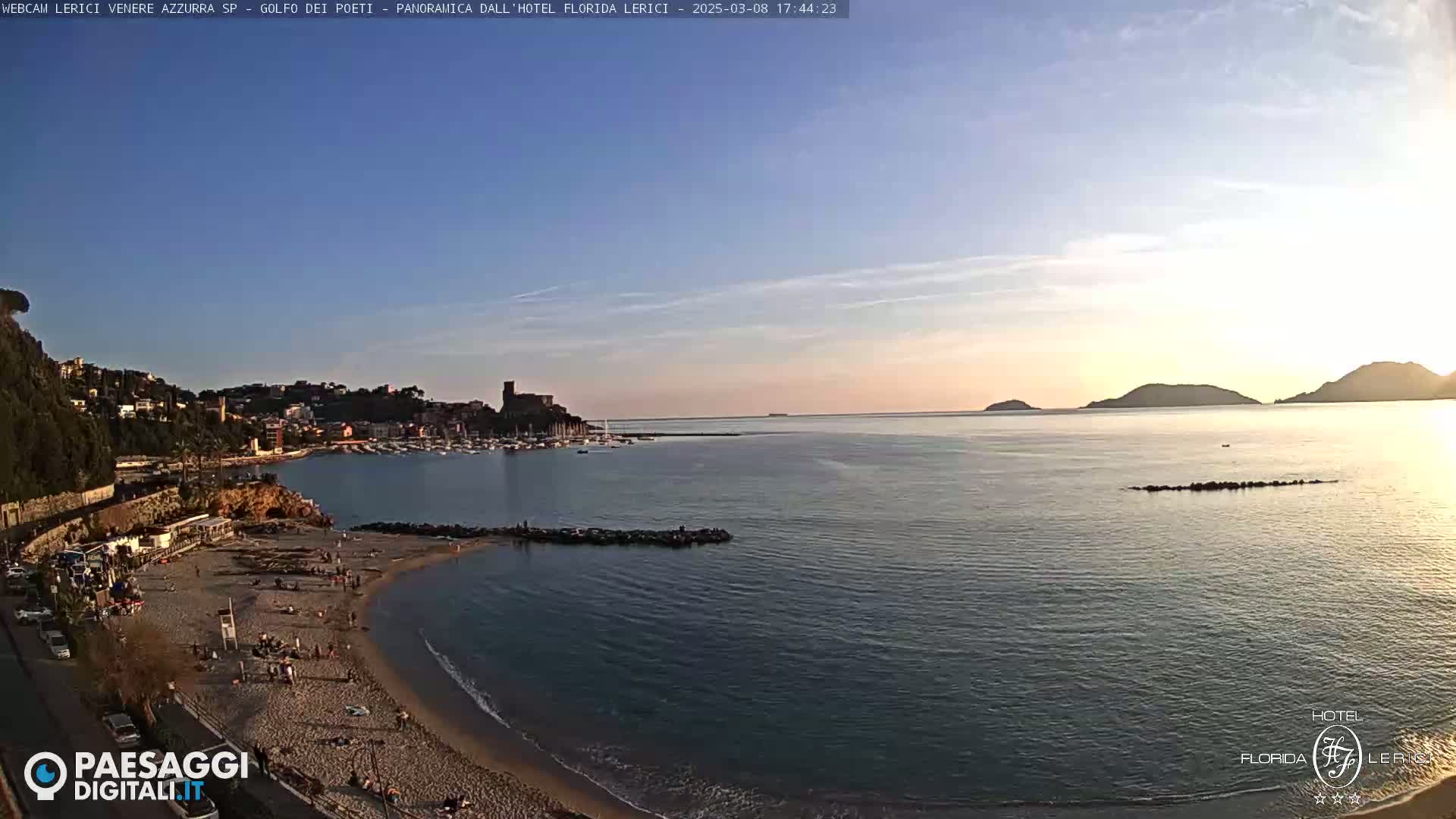 A sandy beach with people on it curves into a calm bay, with a town and hills rising in the background under a clear, sunset sky.