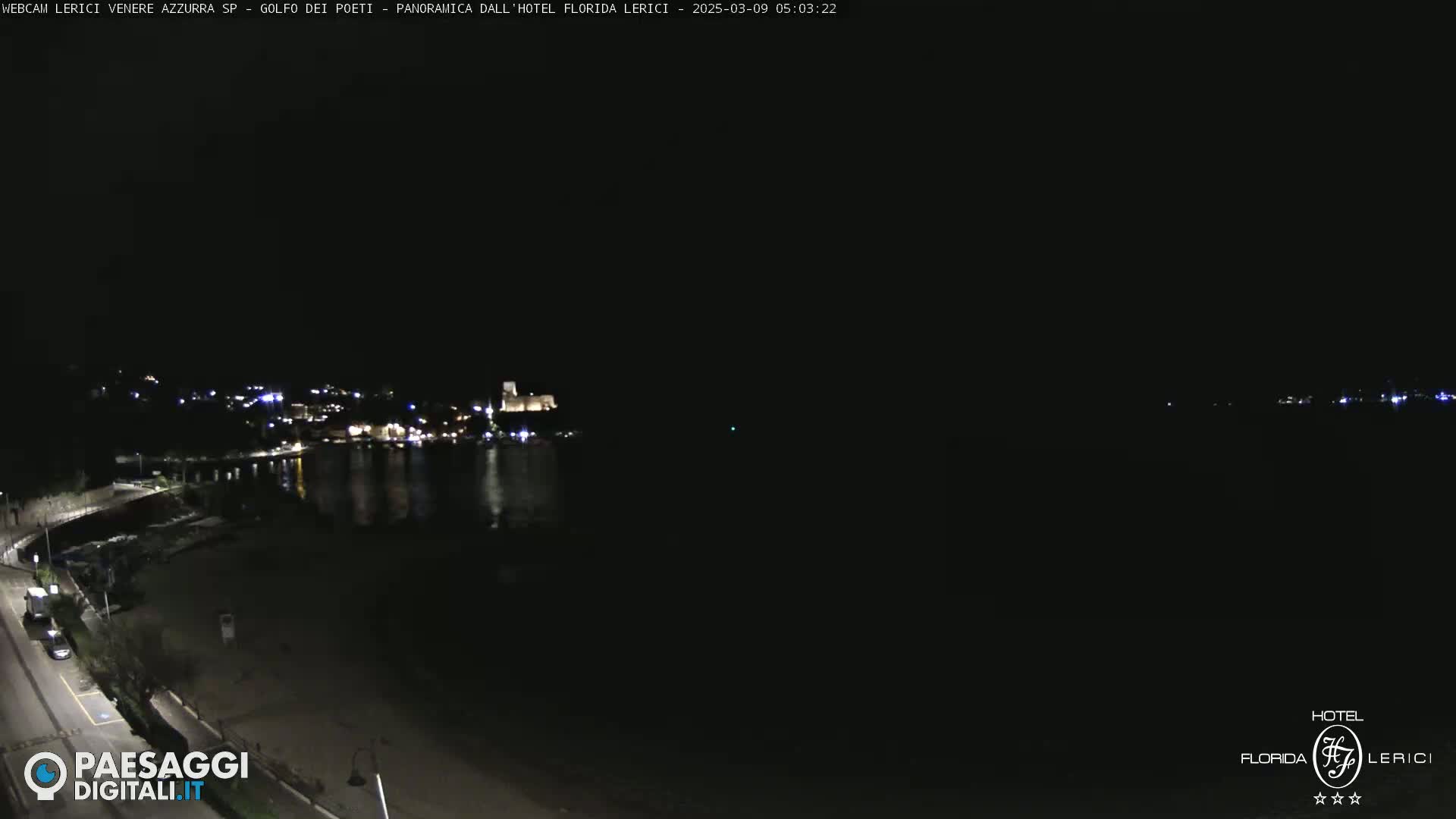 A dark nighttime coastal scene shows a body of water reflecting the lights of a town on the opposite shore, with a mostly empty beach in the foreground.