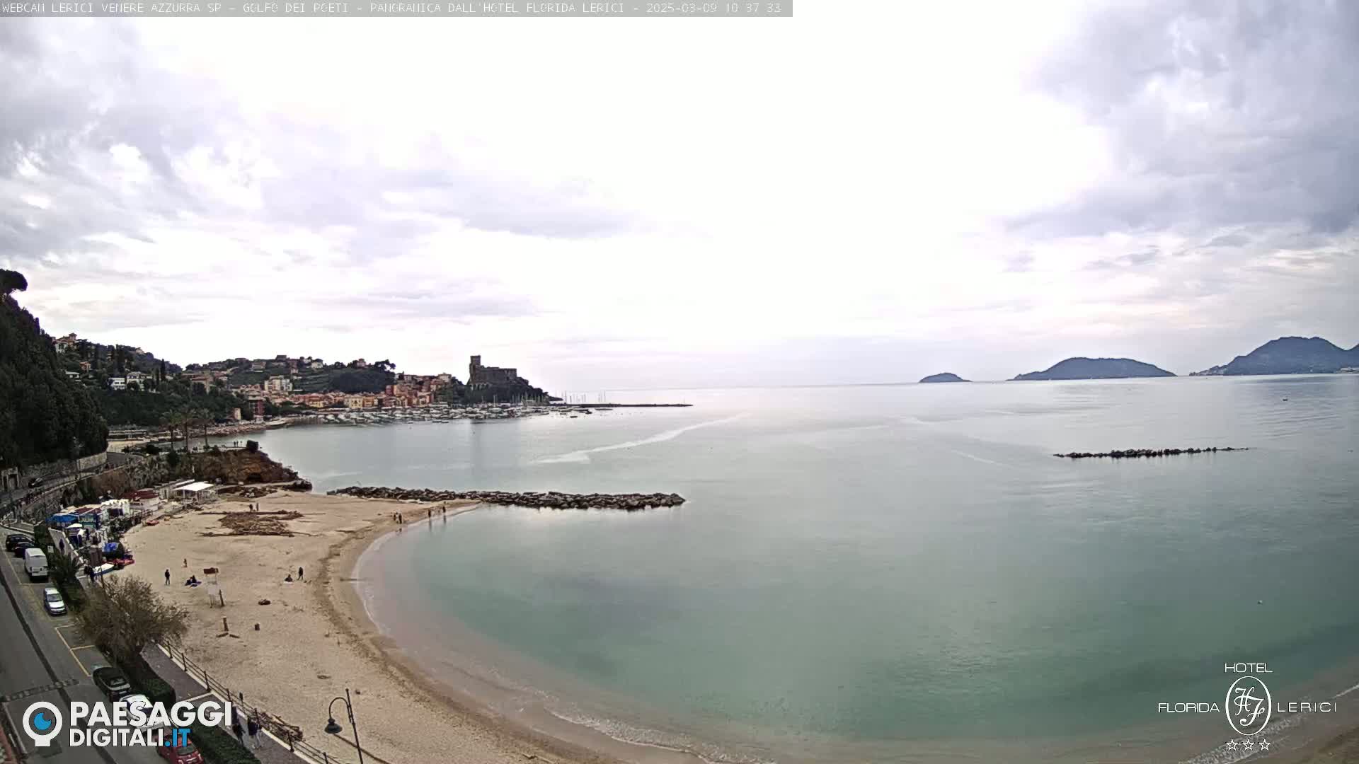 A sandy beach curves into a calm, greenish-blue sea under an overcast sky, with a town and distant hills visible in the background.