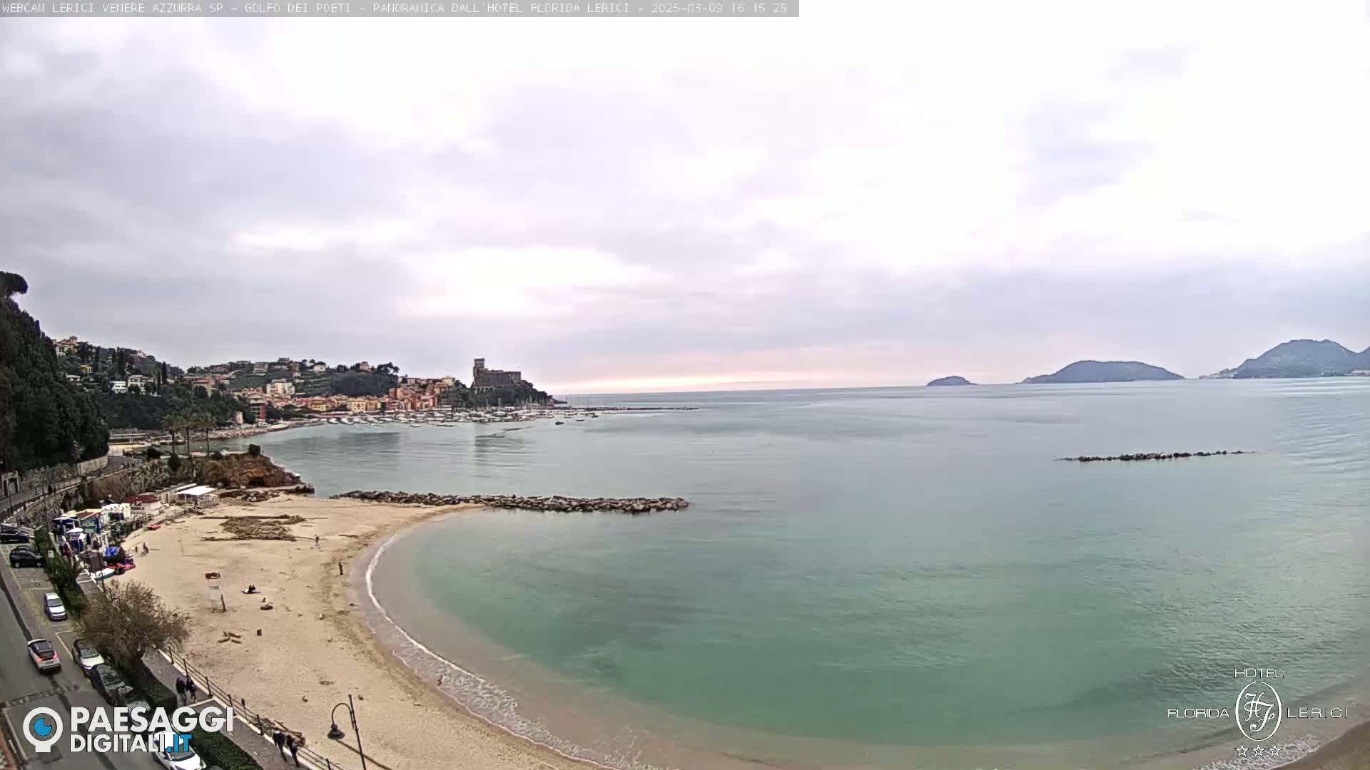 A sandy beach curves into a calm, greenish-blue sea, overlooking a town nestled against hills under a cloudy sky.