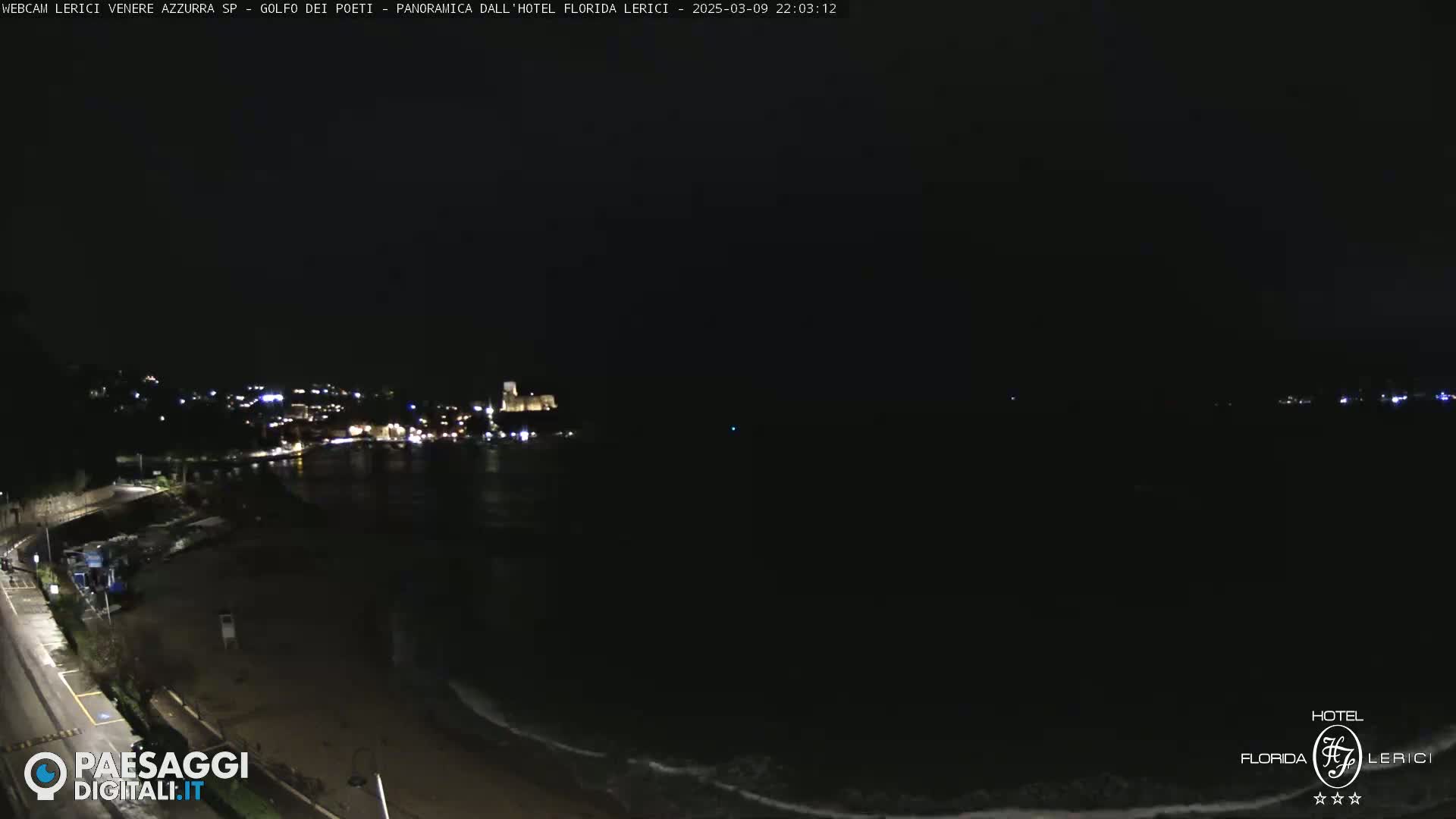 A dark nighttime coastal scene shows a dimly lit town across a calm, dark ocean.