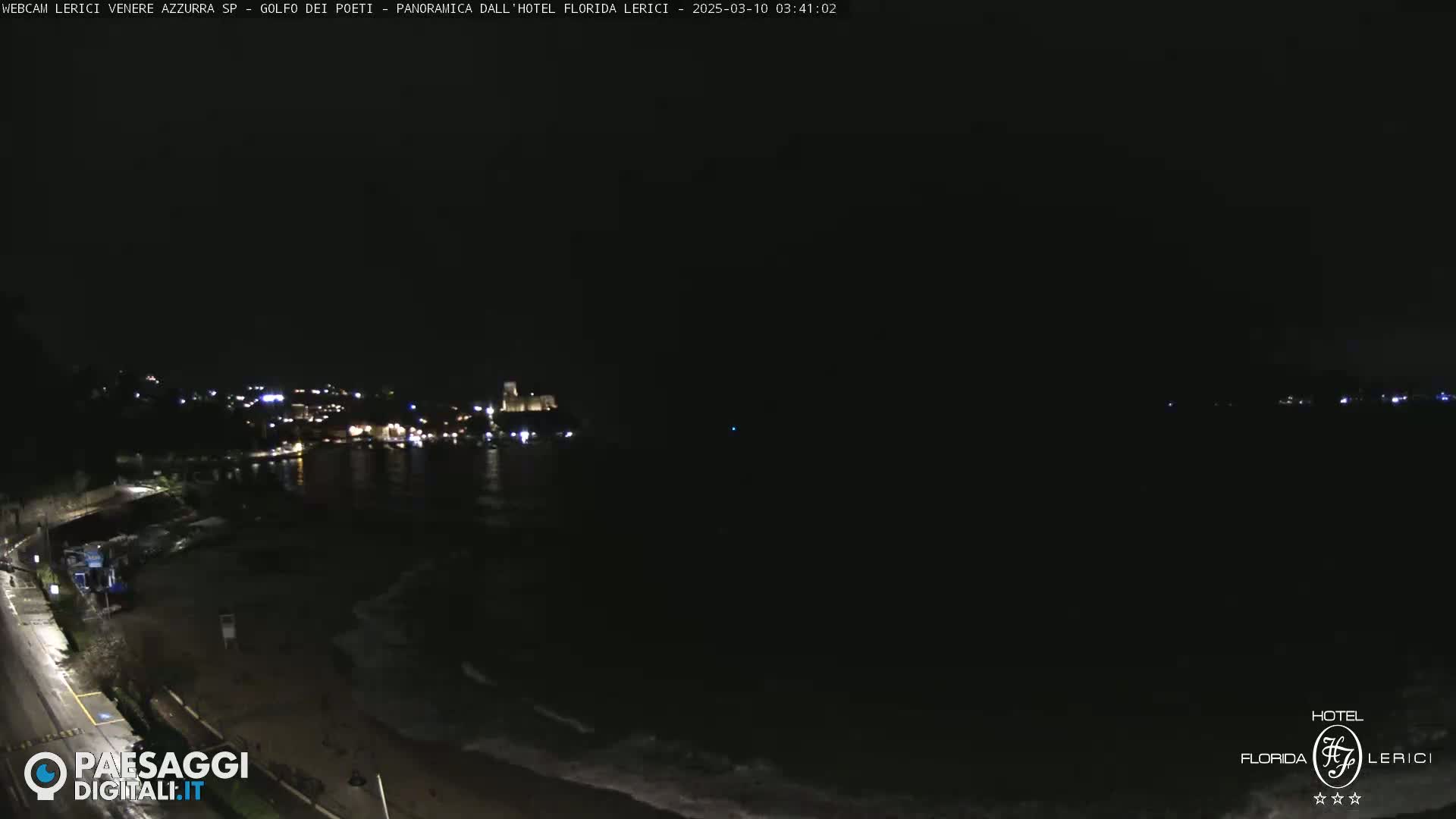 A dark nighttime coastal scene shows a town with lights across a body of water with small waves under a clear, dark sky.