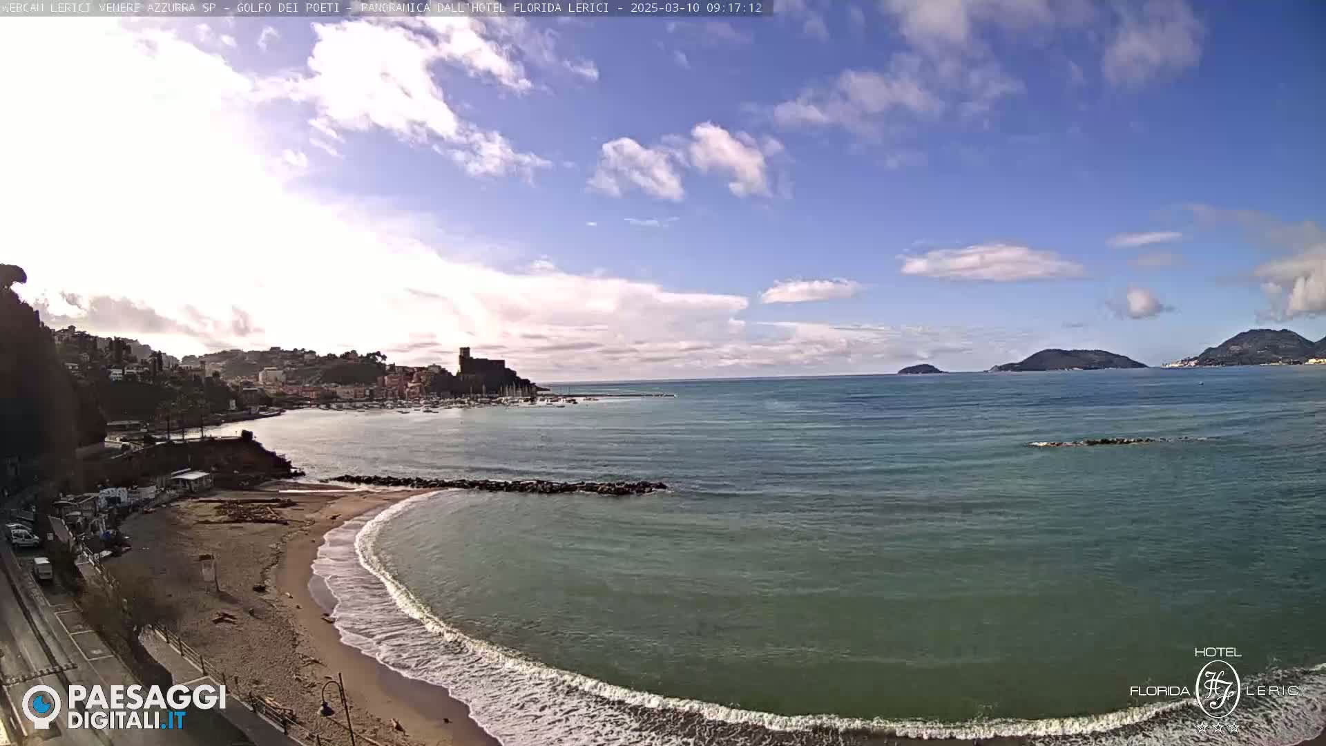 A partly cloudy day shows a sandy beach with small waves lapping the shore, a calm bay with boats, and distant islands under a blue sky.