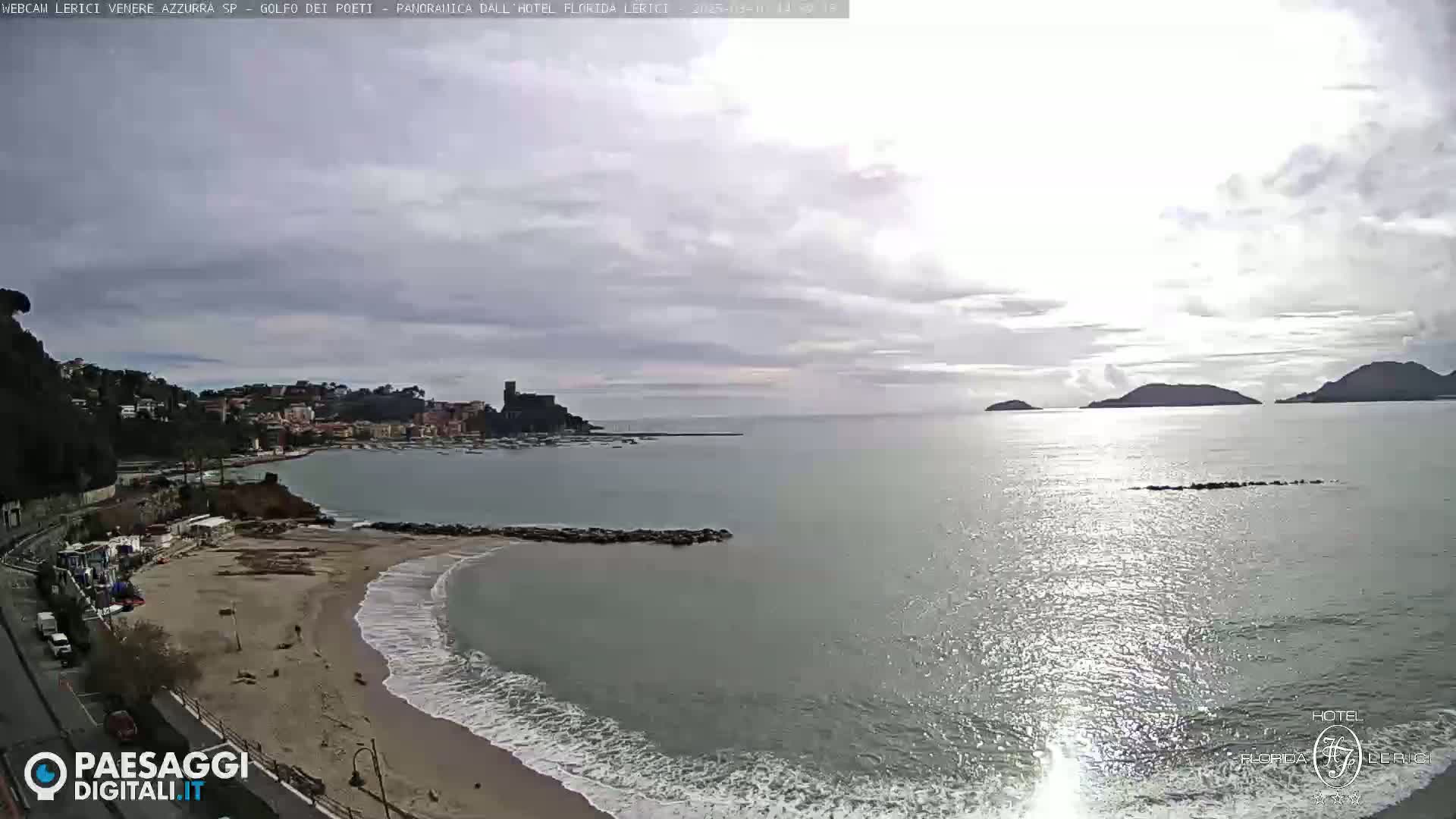 A partly cloudy day shows a sandy beach with waves lapping the shore, a town nestled on a coastline, and islands in the distance.