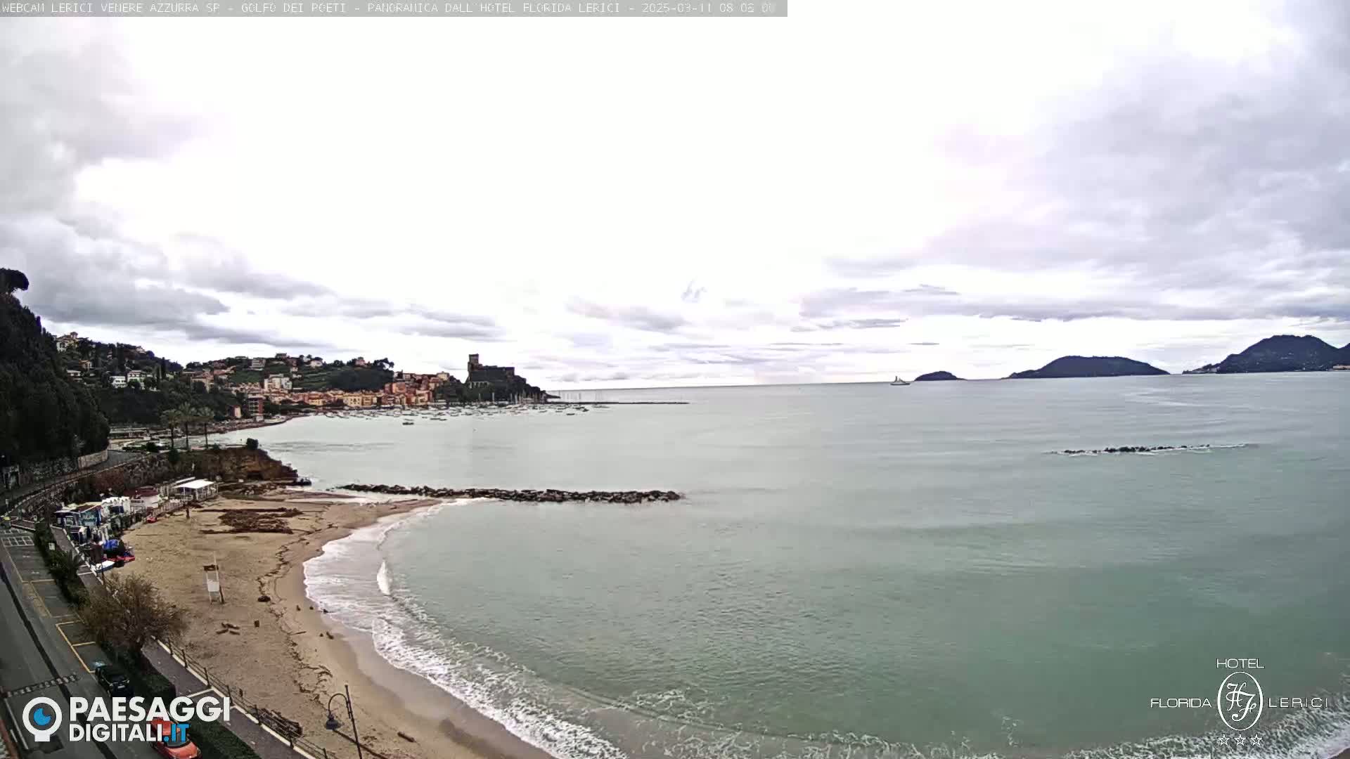 A beach with calm waves laps at the shore, next to a town nestled against a hillside overlooking a bay under a cloudy sky.