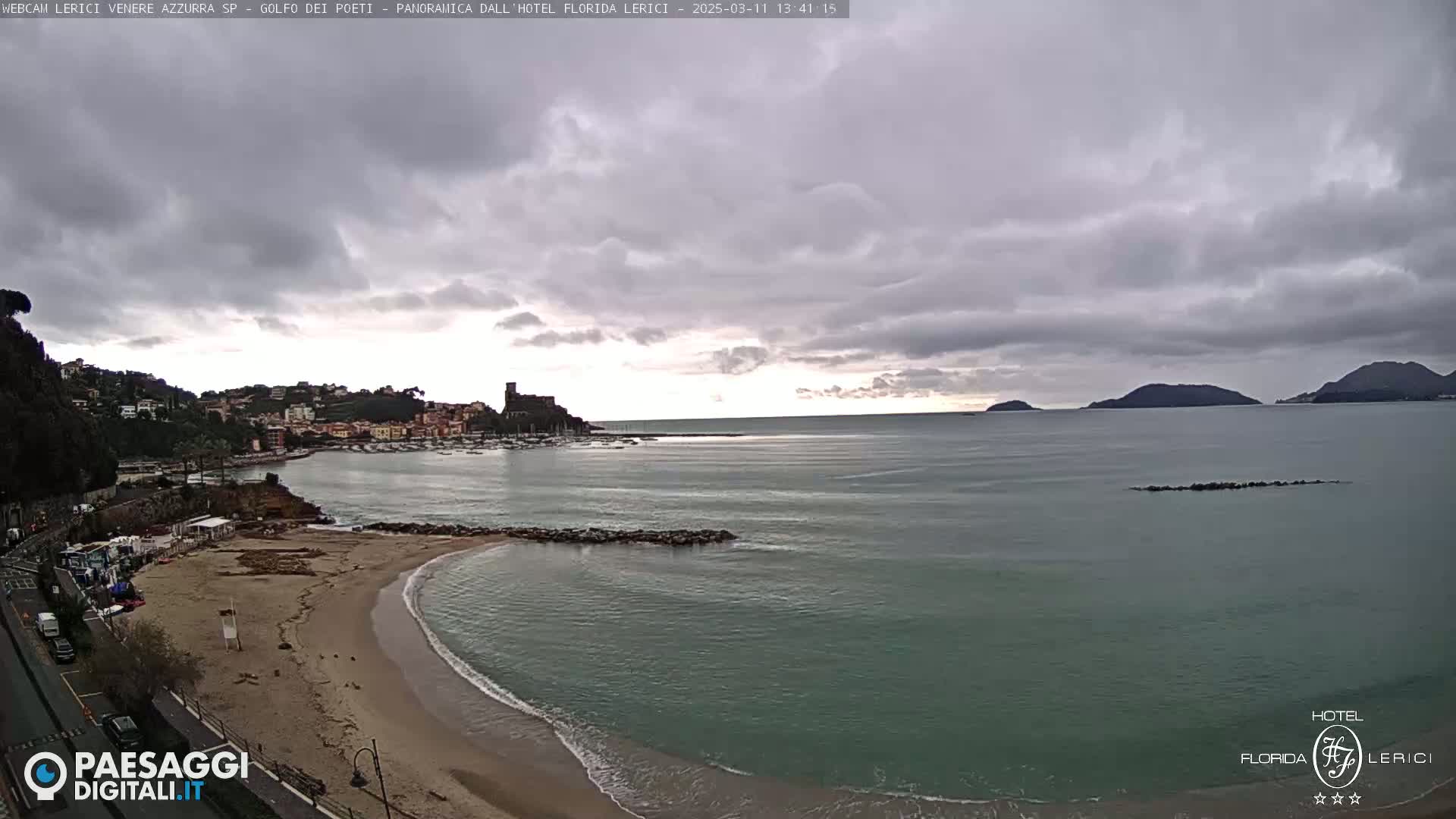 A mostly cloudy day shows a view of a sandy beach curving into a calm bay with a town and distant mountains visible across the water.