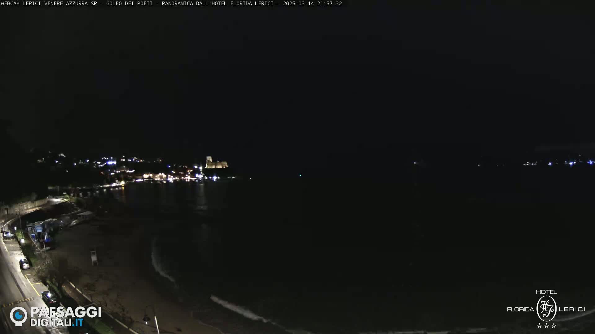A dark nighttime coastal scene shows a dimly lit town across a bay with gentle waves lapping a dark beach.