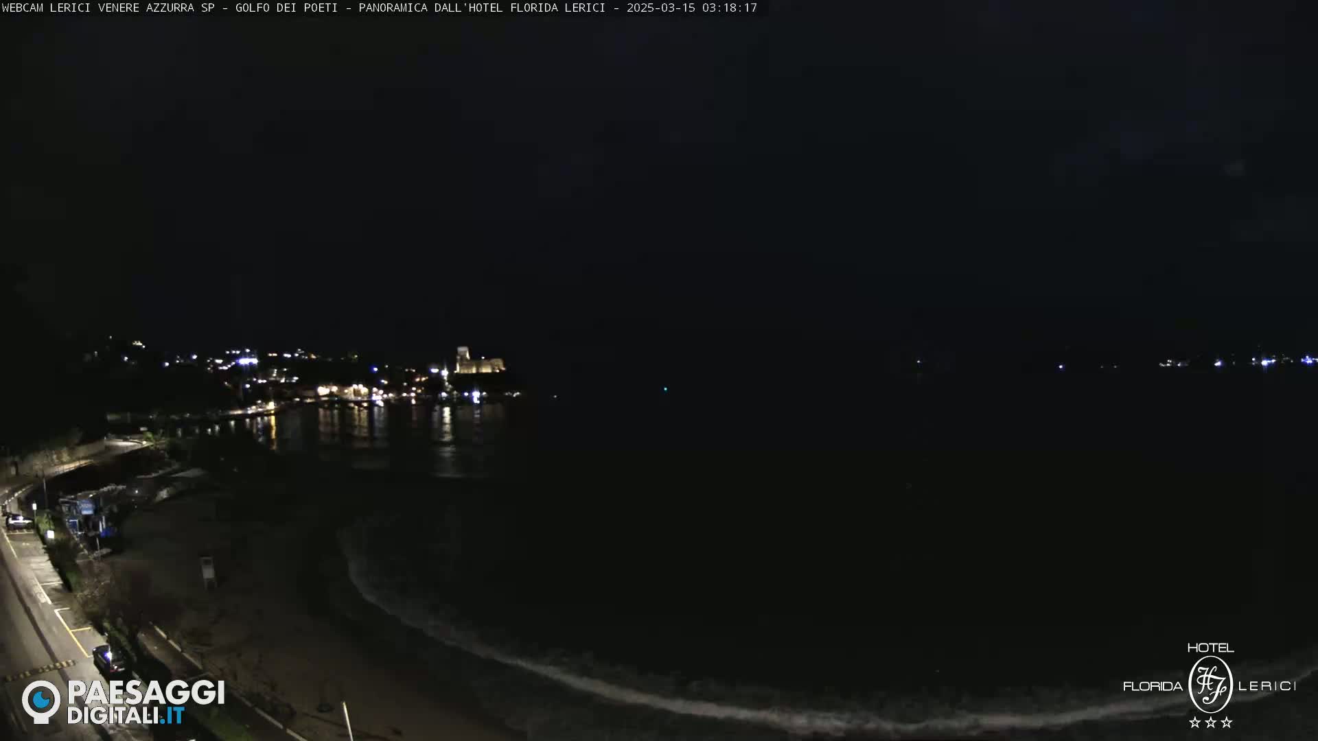 A nighttime coastal scene shows a dark body of water with small waves lapping on a shore, and a dimly lit town in the distance.
