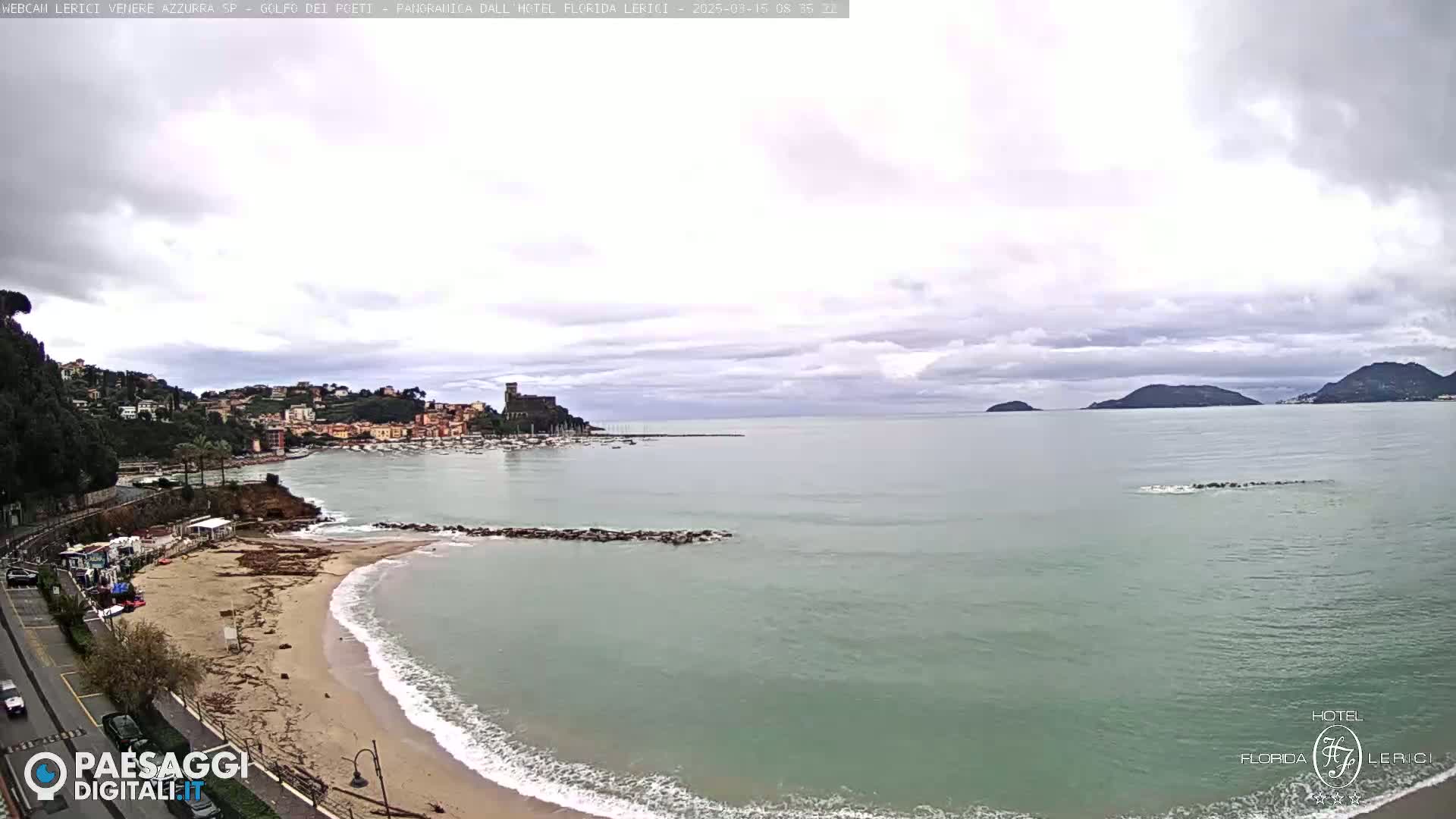 A coastal town with a sandy beach and calm ocean under a cloudy sky is seen from an elevated vantage point.