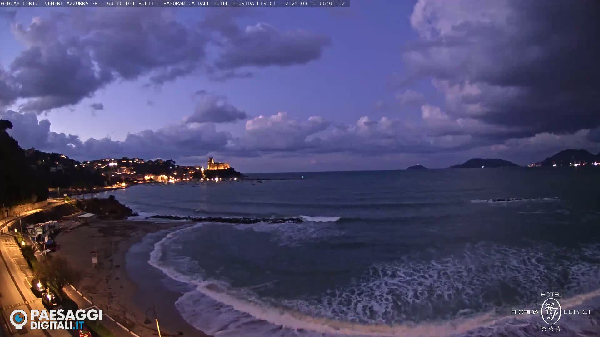 A coastal town at twilight under a partly cloudy sky, with gentle waves lapping a sandy shore, and a castle visible in the distance.