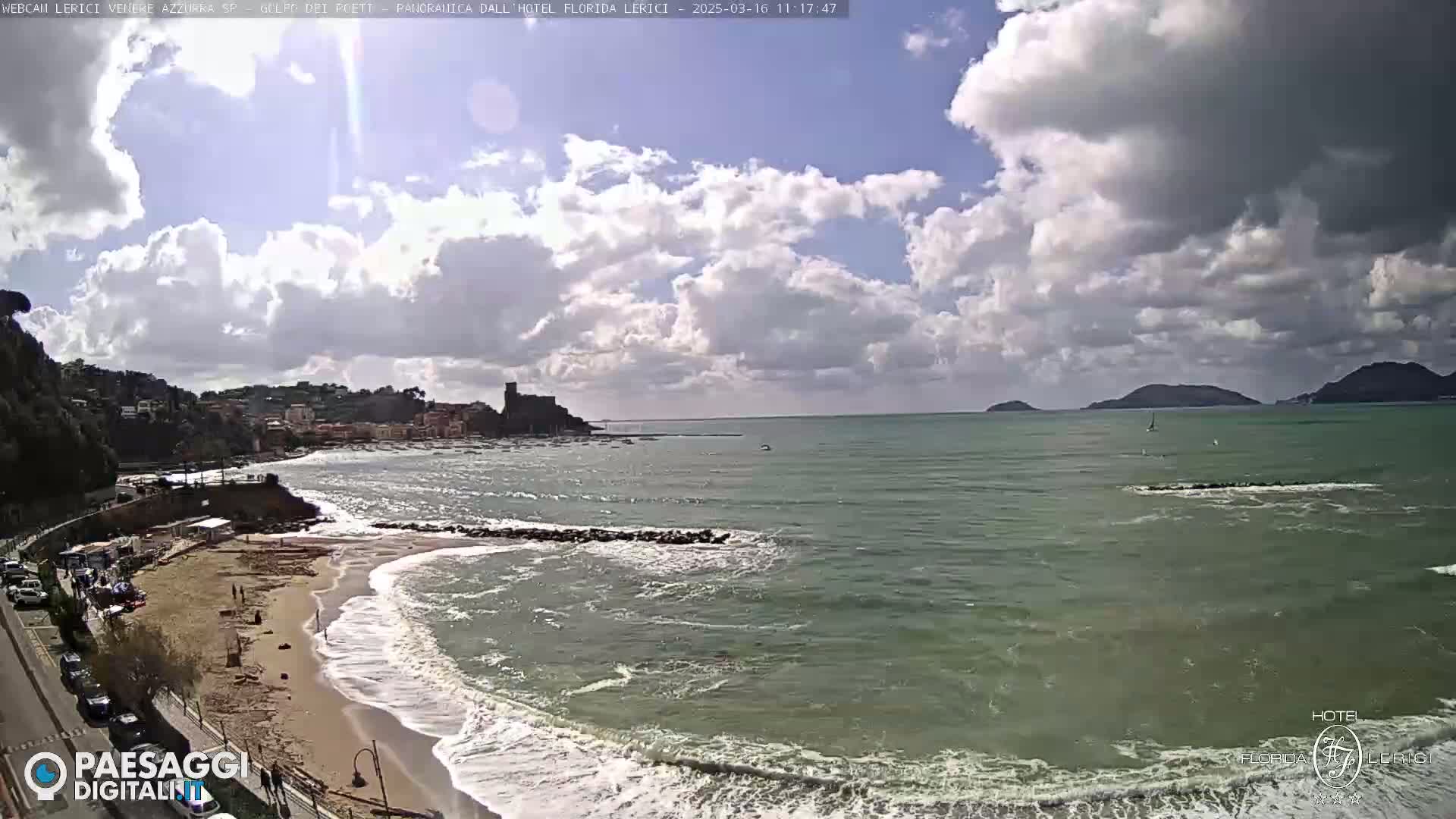 A partly cloudy day shows a sandy beach with waves lapping the shore, a small town nestled against a hillside, and a calm sea extending to distant islands.