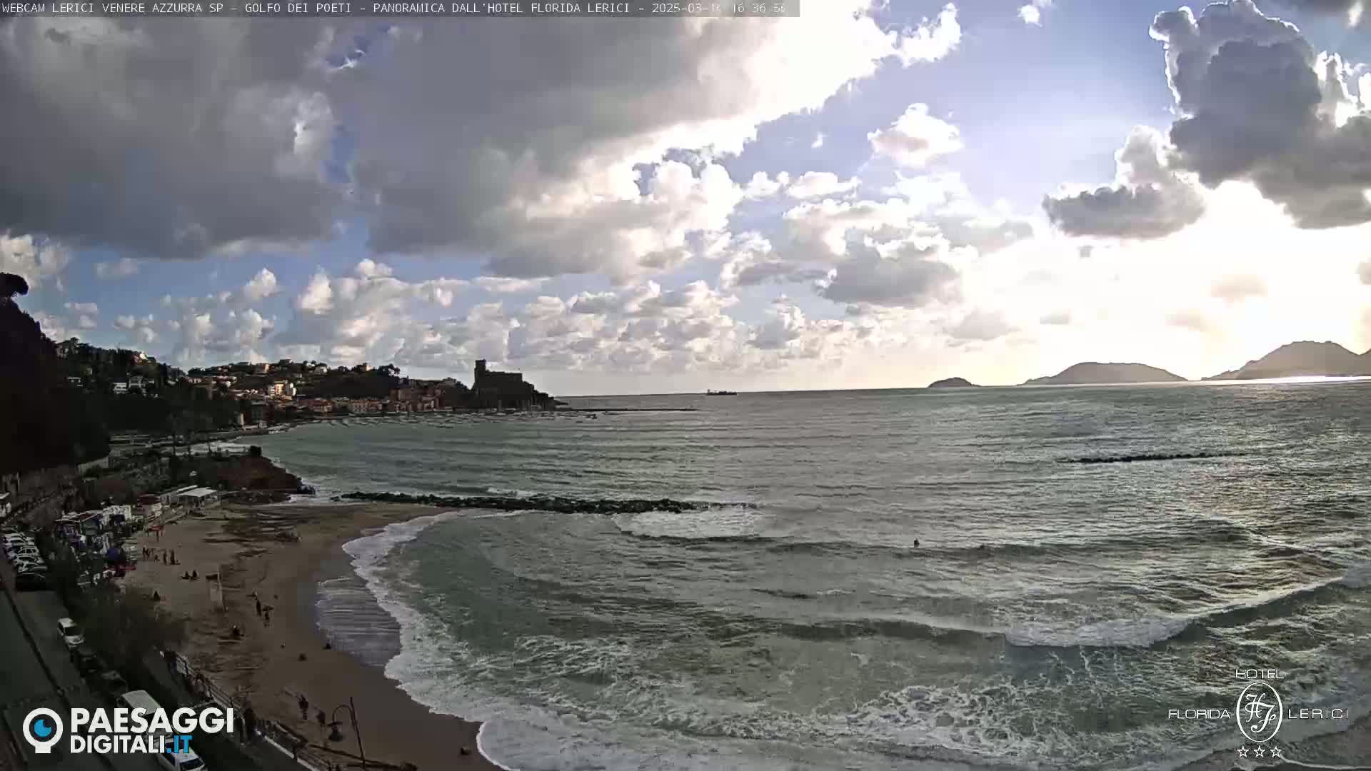 A partly cloudy day shows a coastal town with a beach and ocean waves, hills and islands in the distance.