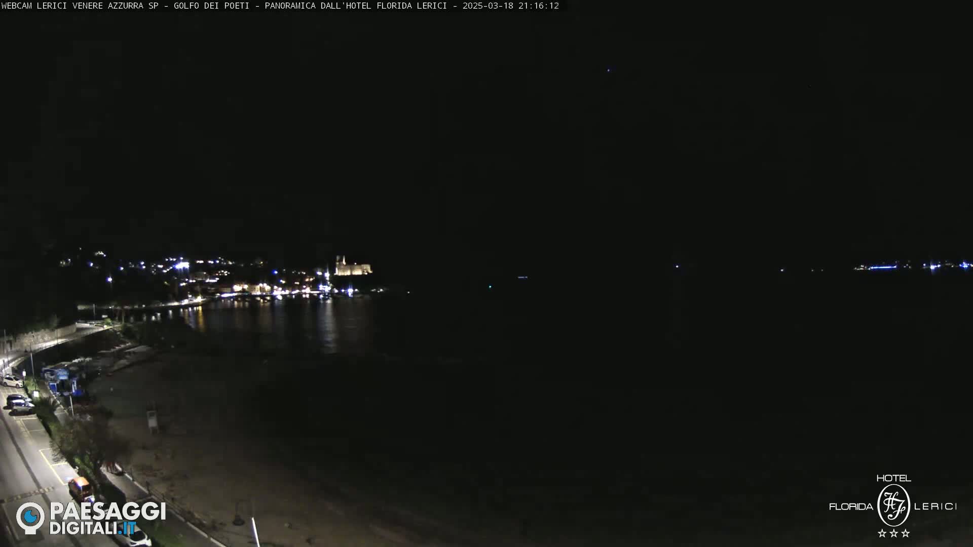 A nighttime view of a dark, calm body of water with a dimly lit town across the water and a beach in the foreground.