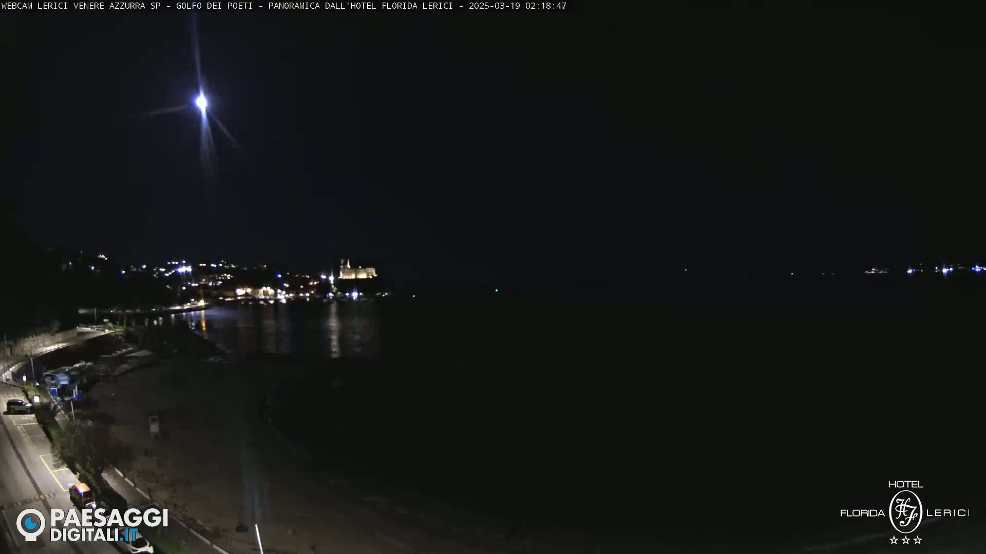 A nighttime coastal scene shows a bright light streaking across the dark sky above a calm body of water and a dimly lit town.