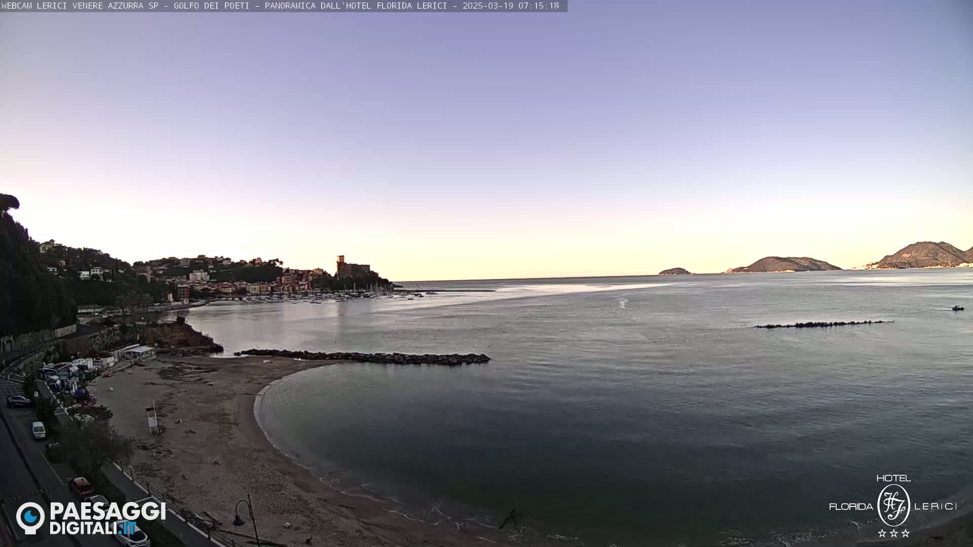A sandy beach curves into a calm bay, with a town and hills visible across the water under a clear, light-blue sky.