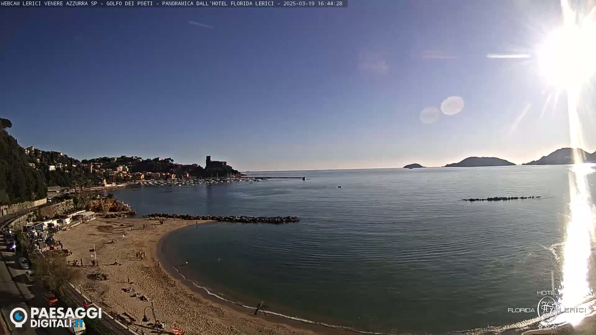 A sunny beach scene shows a sandy cove with people sunbathing, a calm sea with boats in the harbor, and hills in the background.