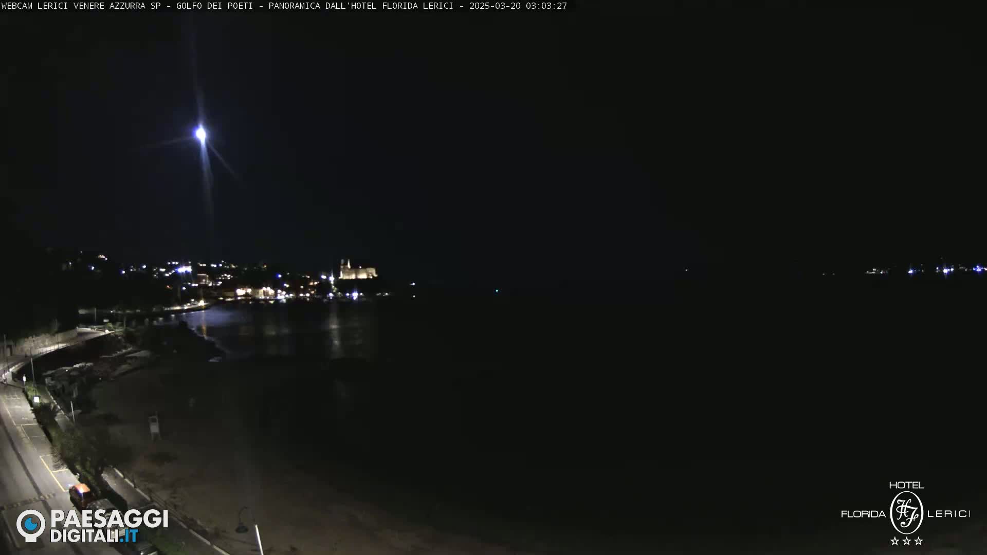 A nighttime, high-angle view shows a dark coastline with a few lights visible, and a bright light streaks across the dark sky above.