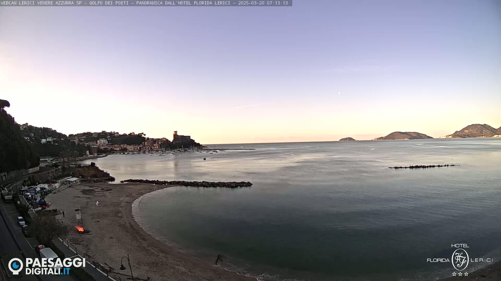 A tranquil coastal scene at dawn shows a sandy beach curving into a calm bay, with a town nestled on a hillside in the background and islands visible on the horizon.