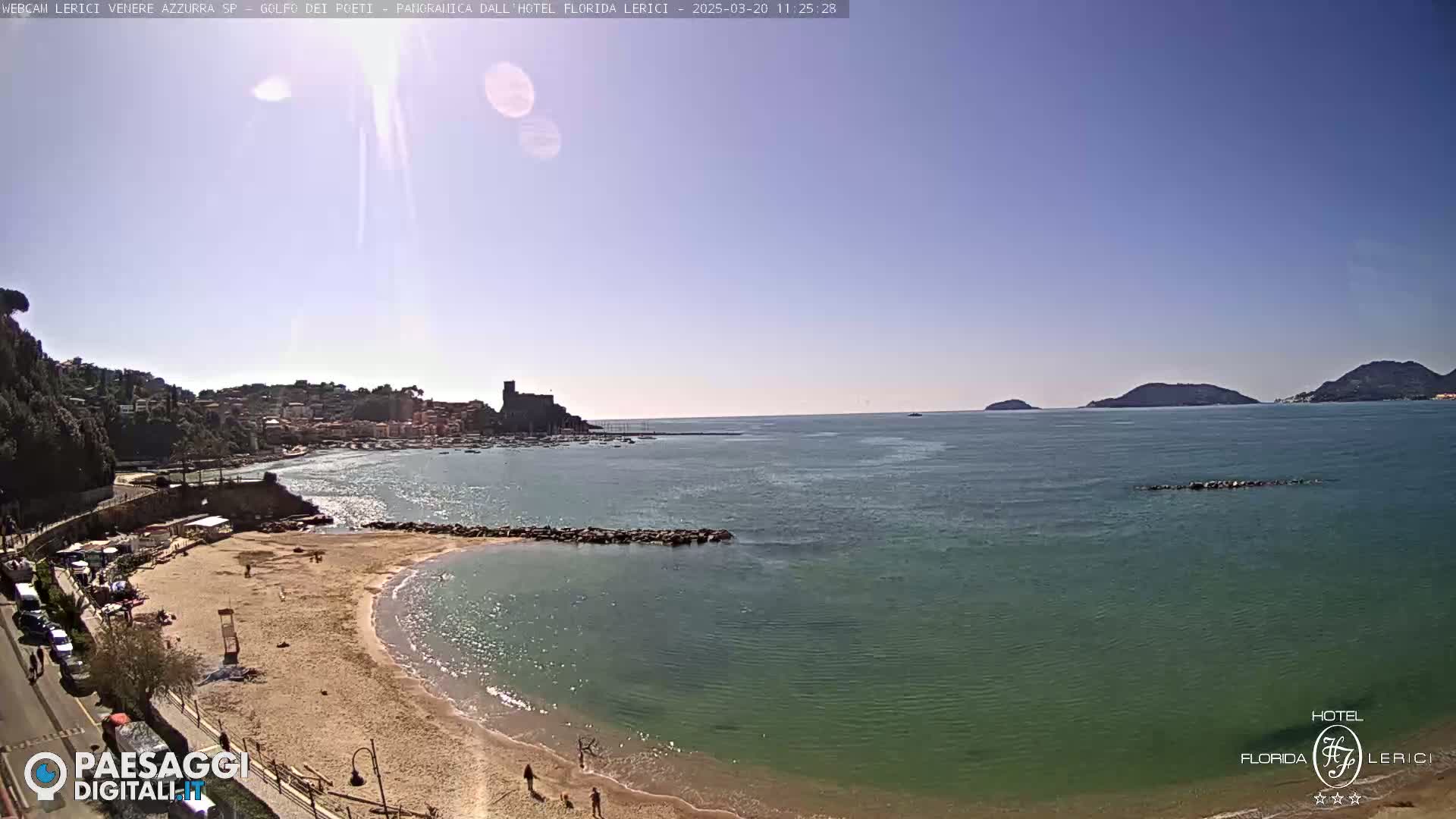 A sandy beach with calm, clear turquoise water stretches toward a town nestled against a hillside, under a clear blue sky.