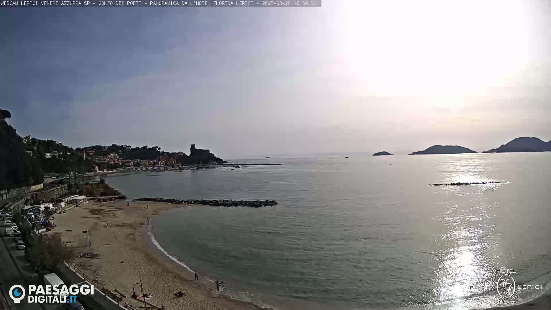 A mostly sunny day shows a sandy beach curving into a calm bay, with a town and distant islands visible across the water.
