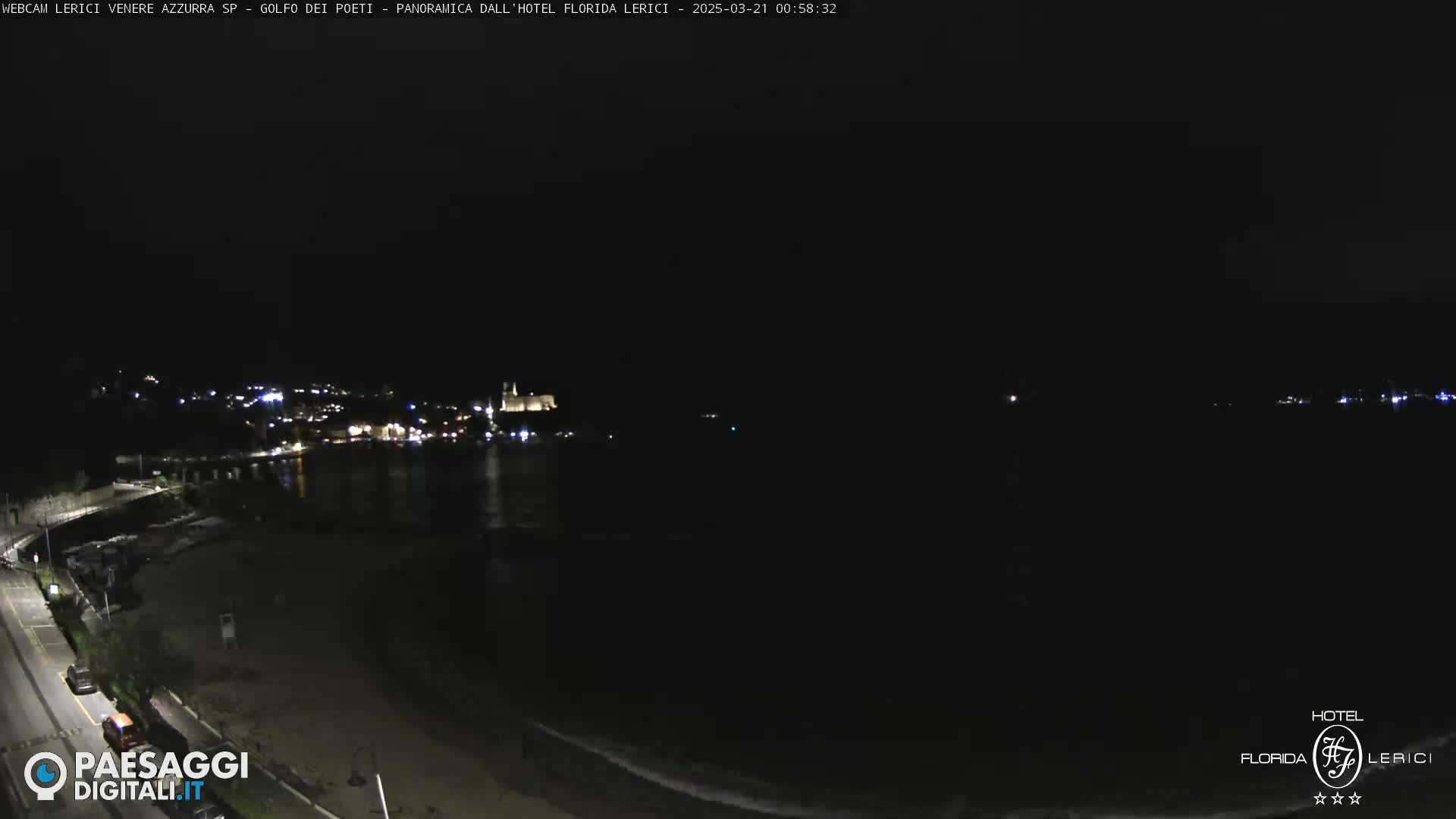 A nighttime view of a dark beach and calm water, with a town's lights visible in the distance across the water.