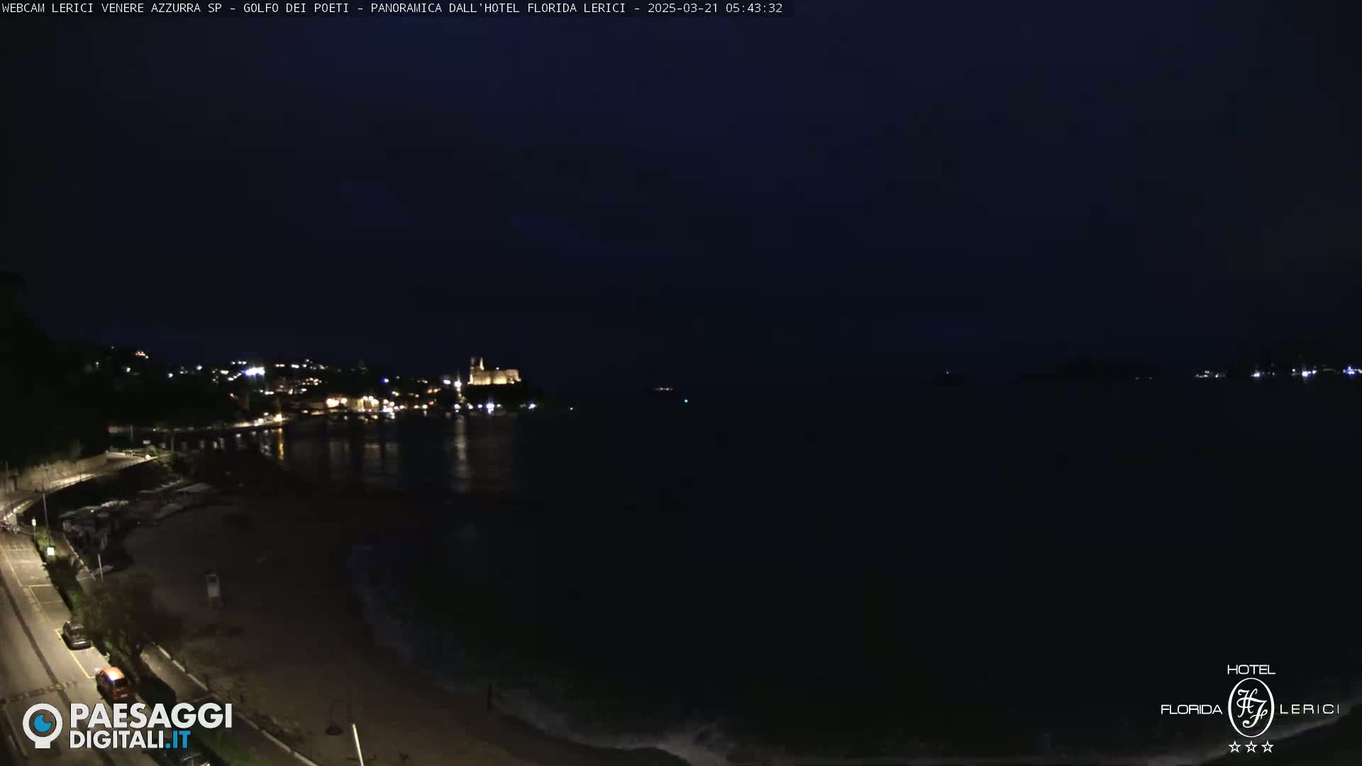 A nighttime coastal scene shows a dark body of water with dimly lit buildings and a road along the shoreline.