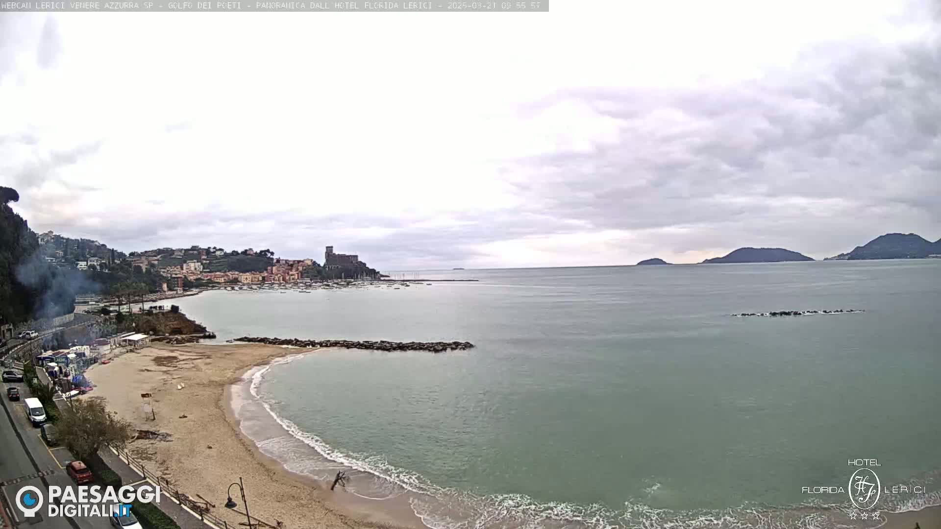 A sandy beach with calm waves laps the shore near a town nestled against hills, all under a cloudy sky.