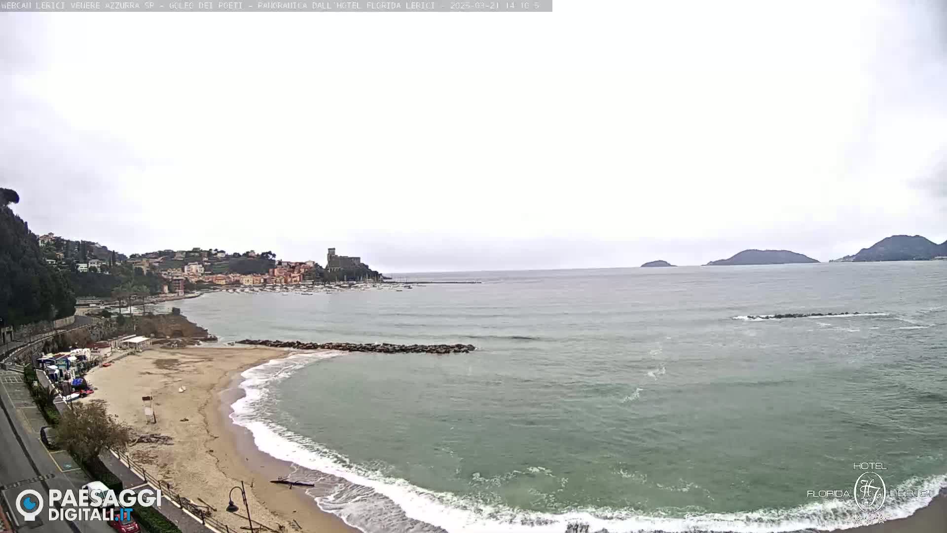 A sandy beach with gentle waves laps the shore, overlooking a calm bay with a town and distant hills under an overcast sky.