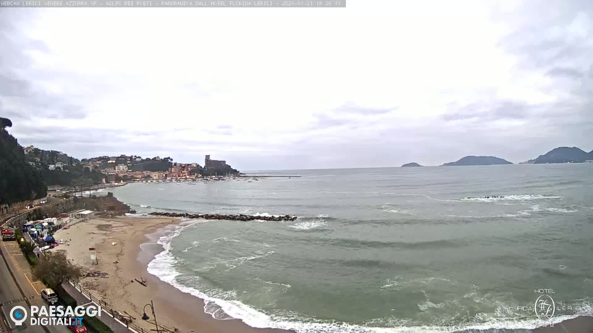 A coastal scene with overcast skies shows a sandy beach with waves lapping the shore, a town nestled against hills, and calm water extending to distant islands.