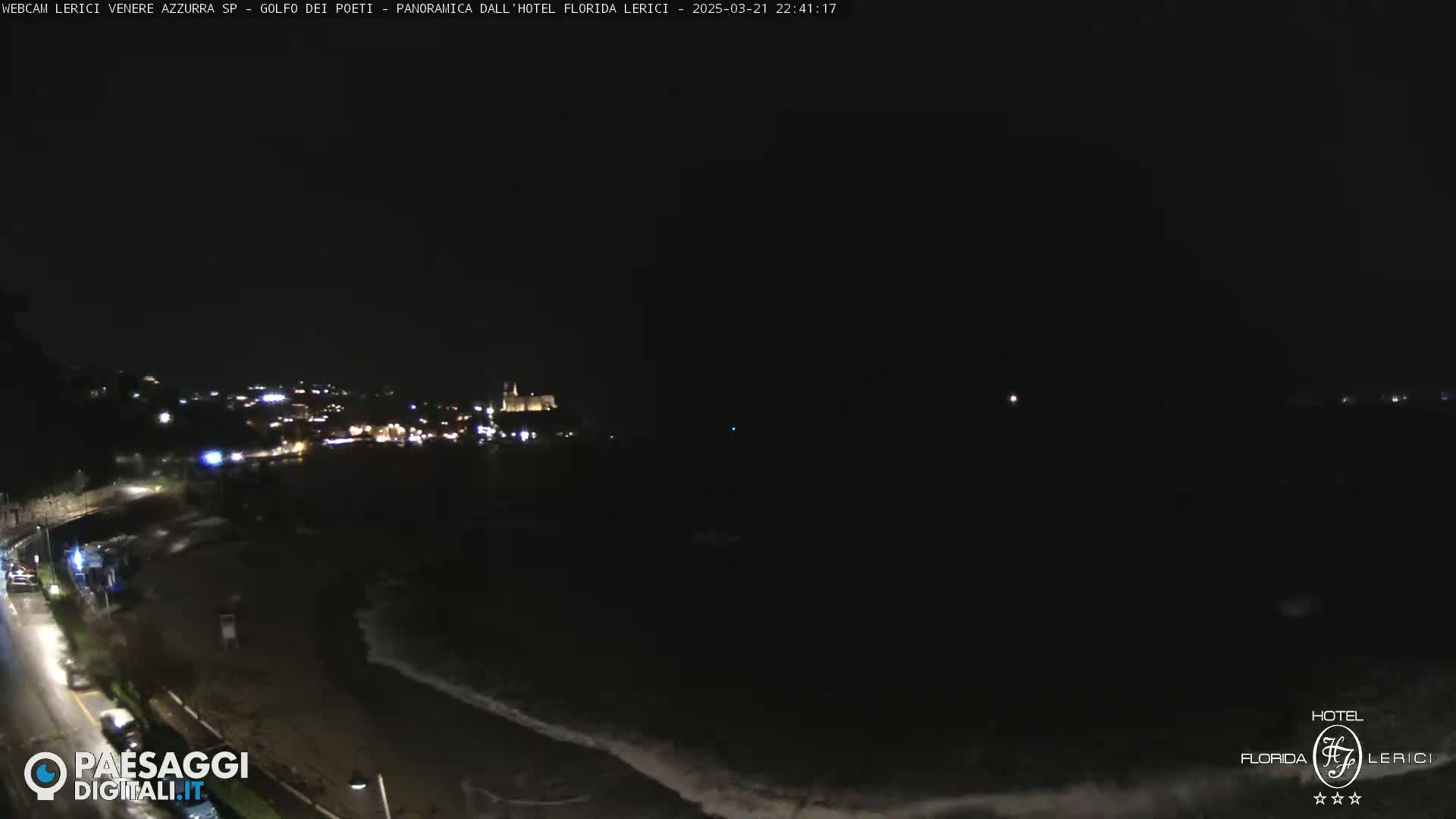 A nighttime coastal scene shows a dark ocean with small waves lapping on the shore, a dimly lit town in the distance, and a road with a few cars visible.