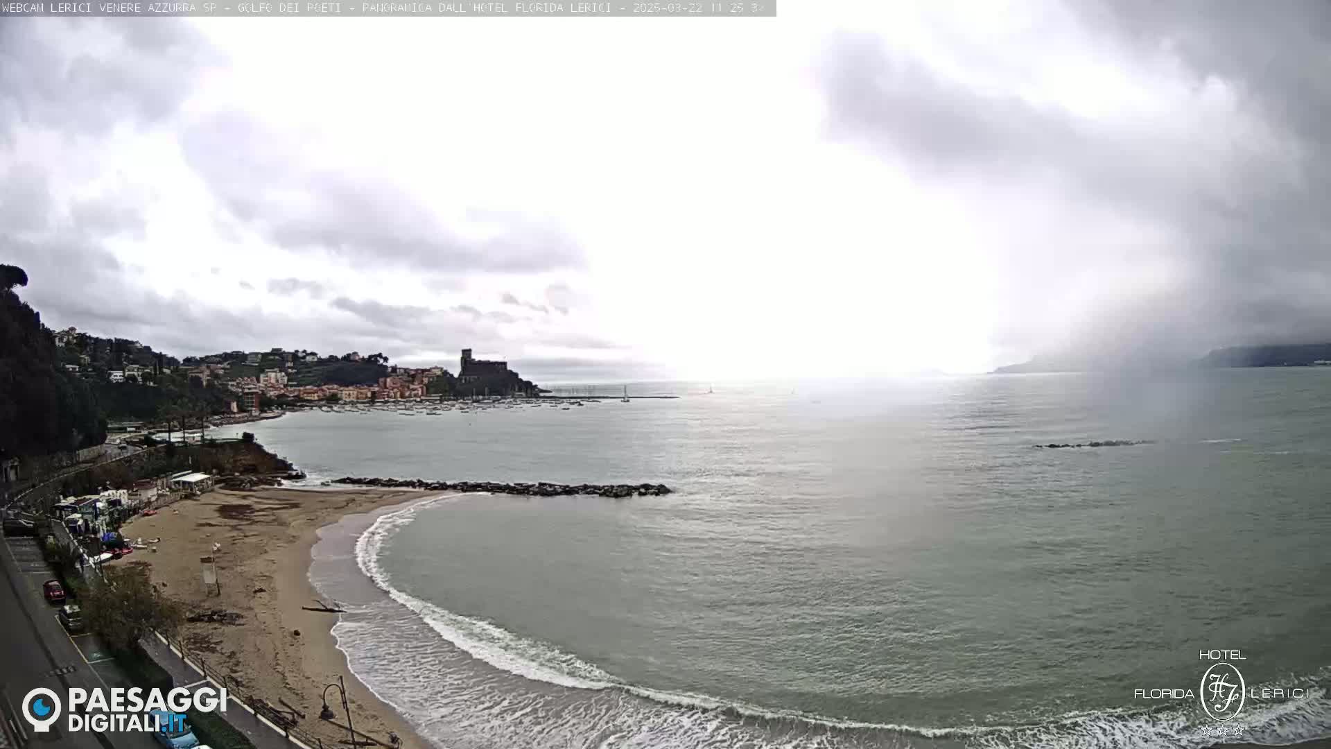 A coastal town with a sandy beach and calm waves is seen under an overcast sky.