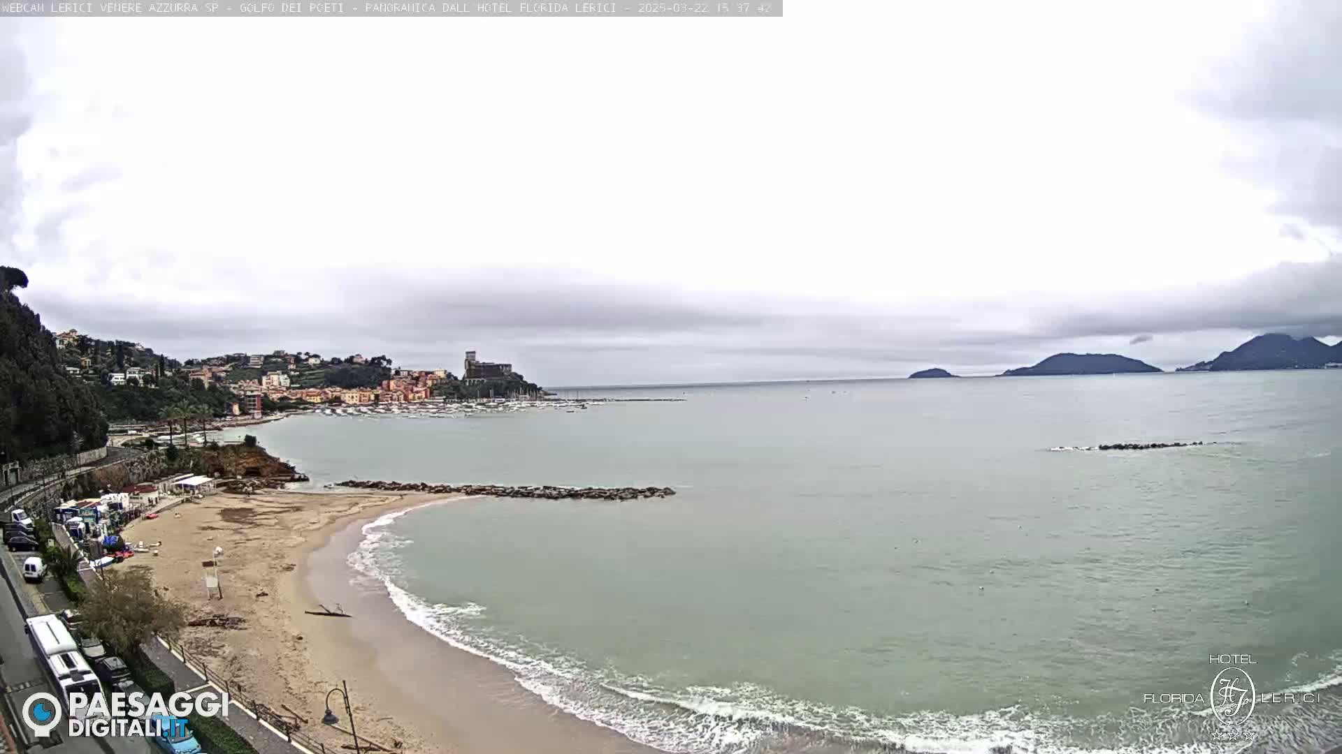 A sandy beach with calm, light waves laps the shore of a bay, overlooked by a town nestled on a hillside under an overcast sky.