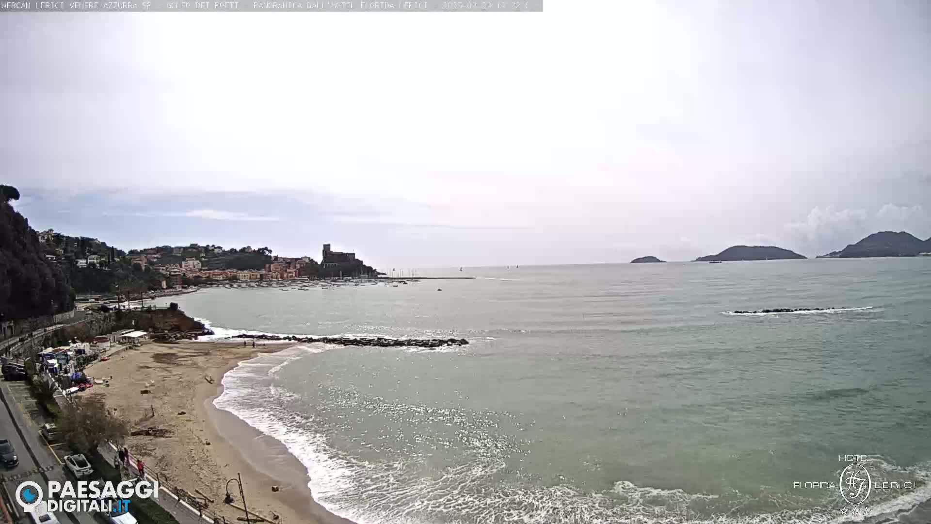 A sandy beach with gentle waves laps against the shore, in front of a town nestled amongst hills, with a calm sea extending to distant islands under an overcast sky.