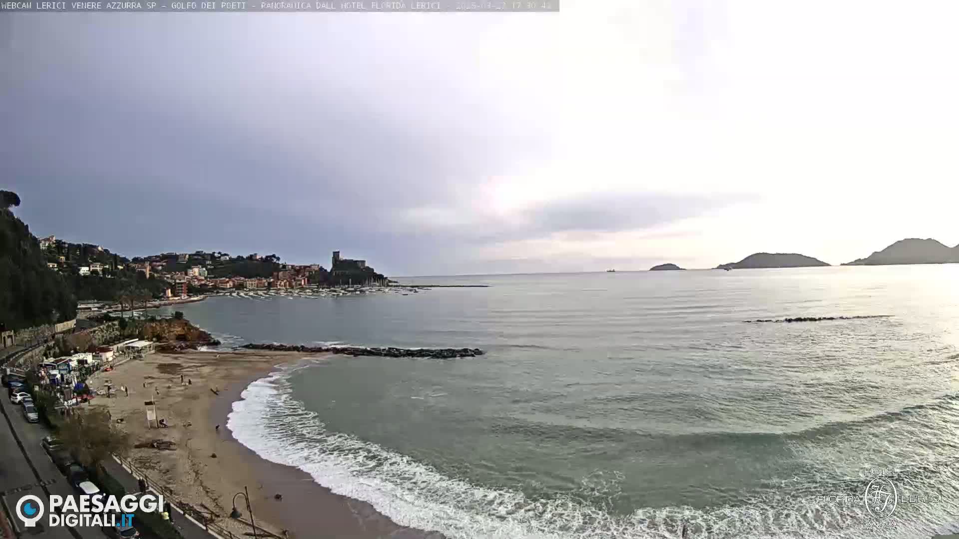 A beach with gentle waves laps at a sandy shore, in front of a calm sea and a town nestled against a hillside under a cloudy sky.