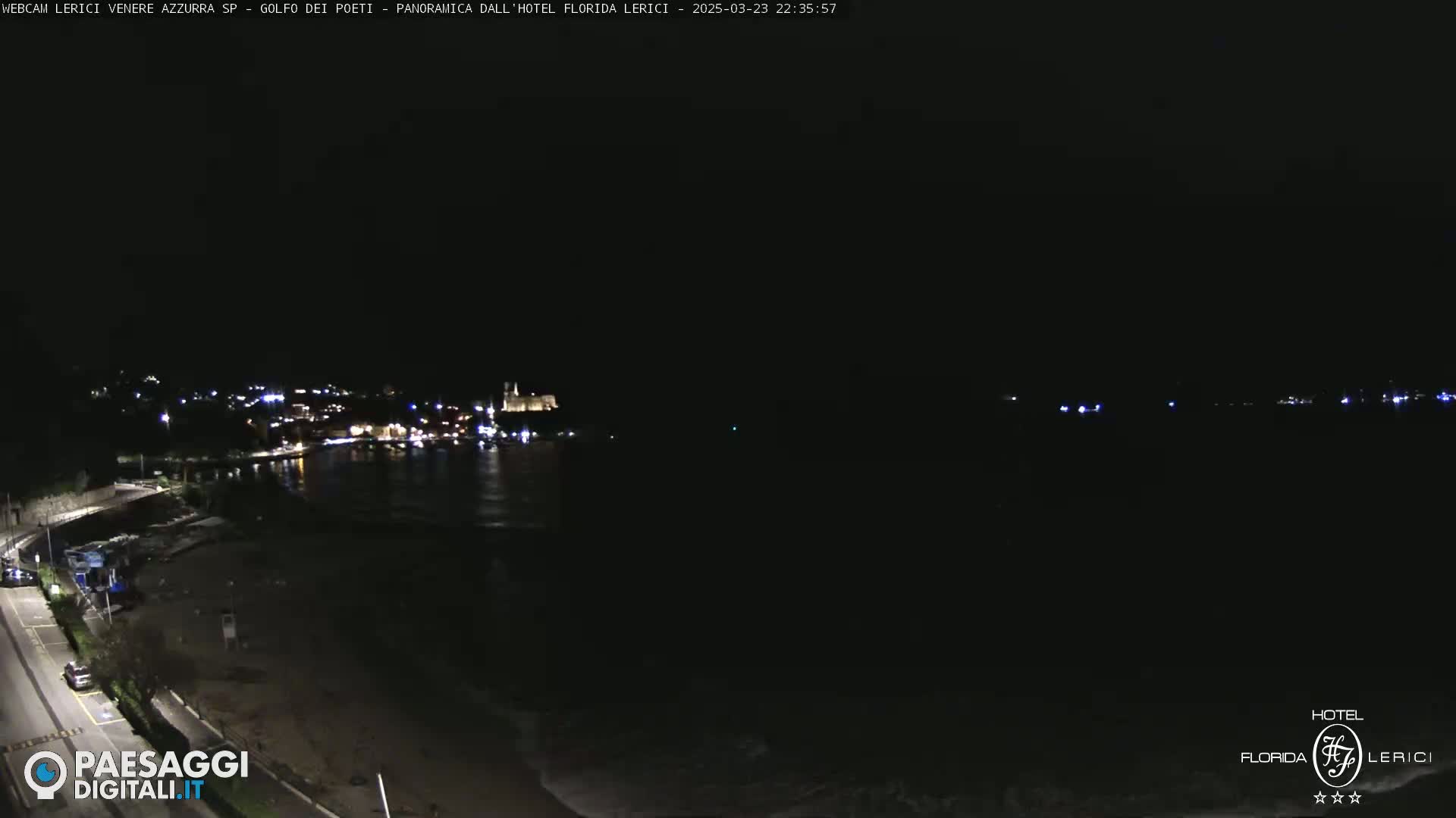A nighttime coastal scene shows a dark beach and calm water reflecting the lights of a town across the bay.