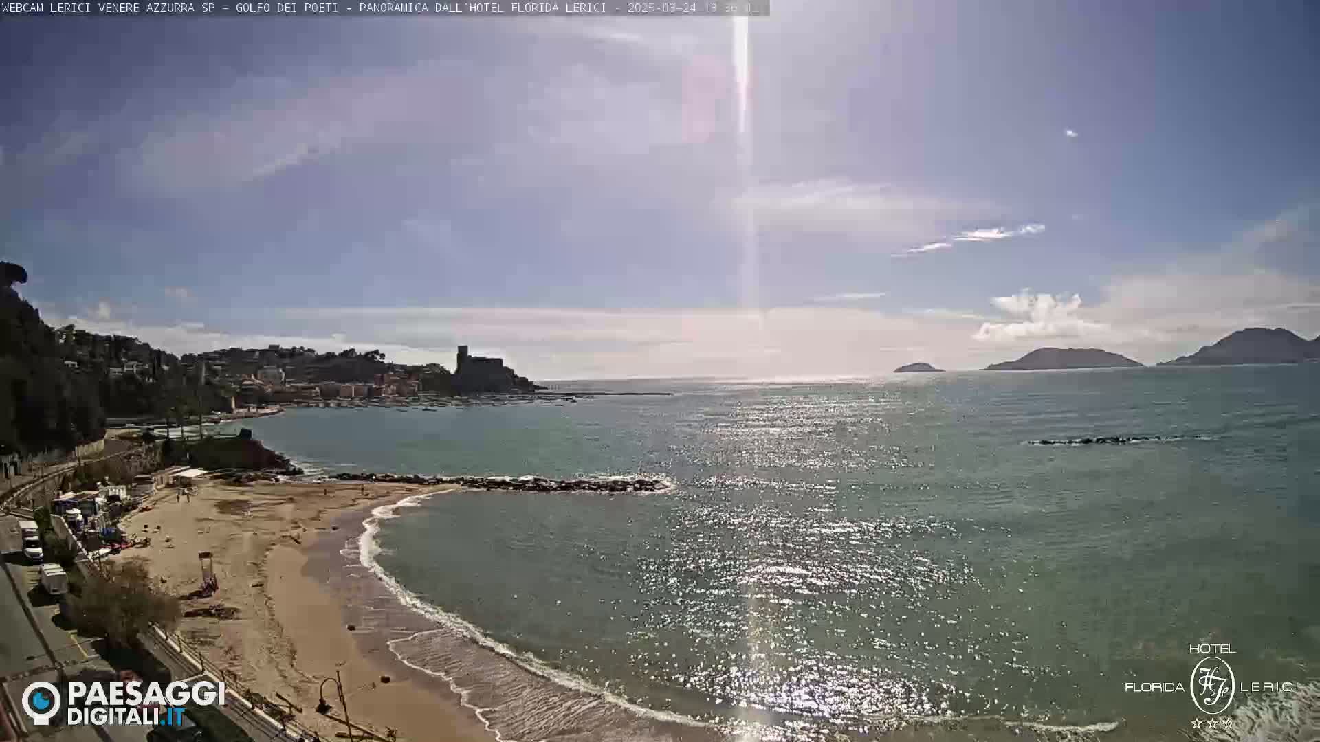 A sandy beach with calm, sparkling water stretches towards a distant coastline under a mostly sunny sky with a few scattered clouds.