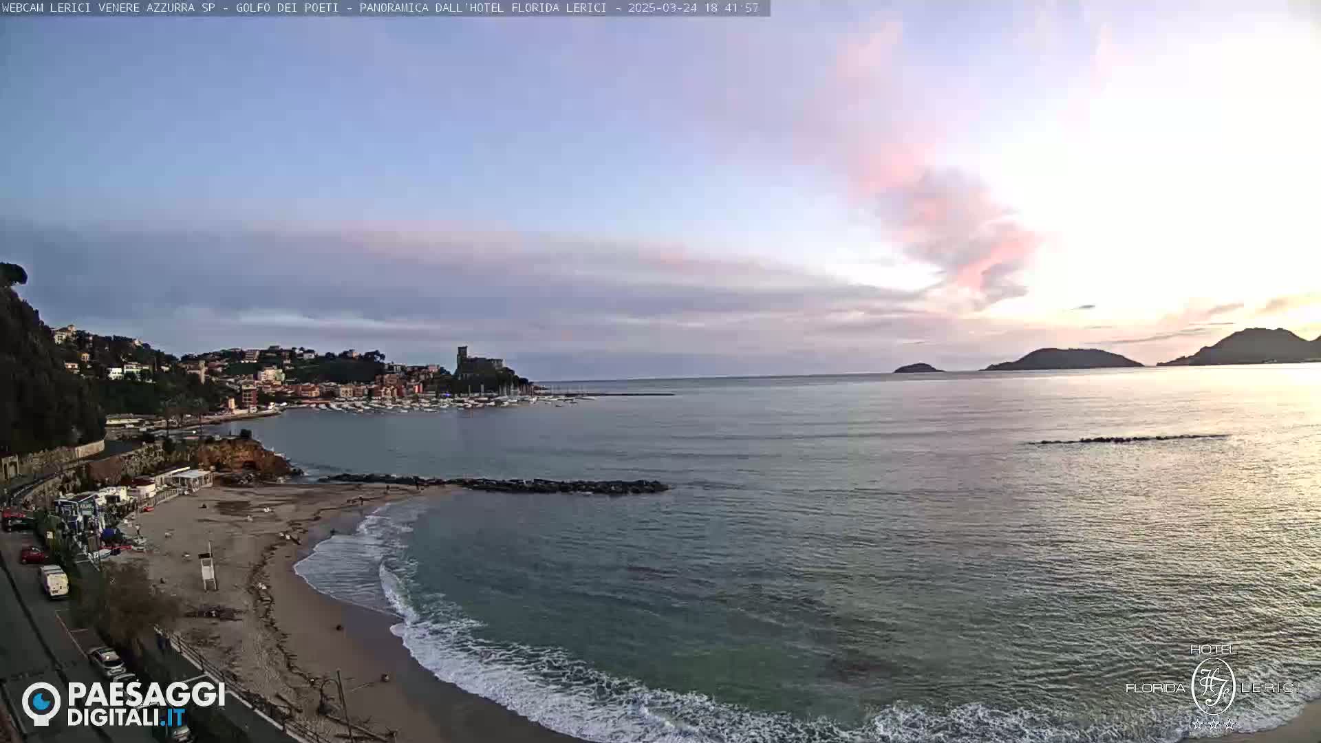 A sandy beach with gentle waves laps the shore under a partly cloudy twilight sky, overlooking a calm bay with a town nestled on a hillside and distant mountains.