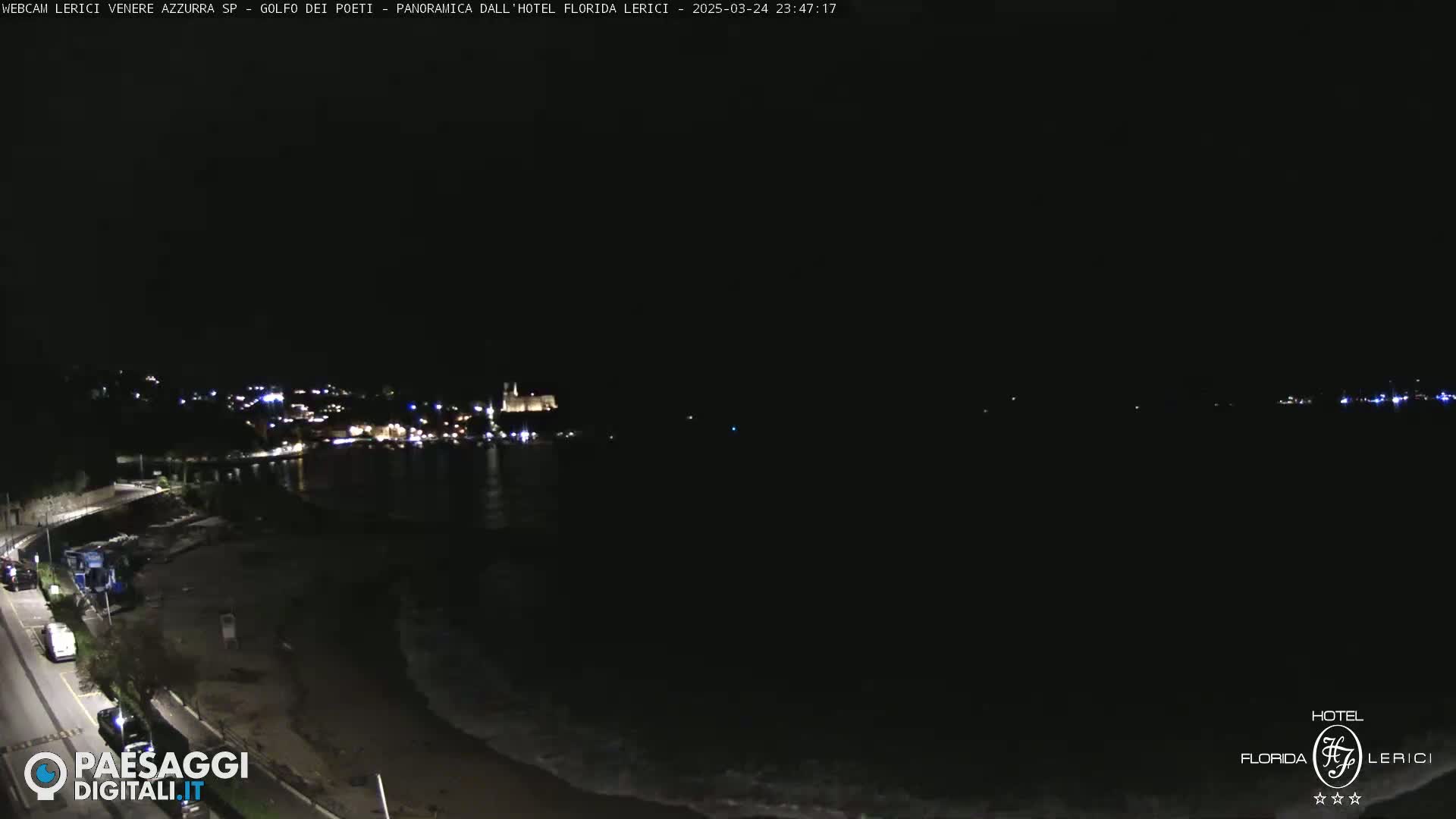 A nighttime view of a dark coastline with a town's lights visible in the distance, and gentle waves lapping the shore.