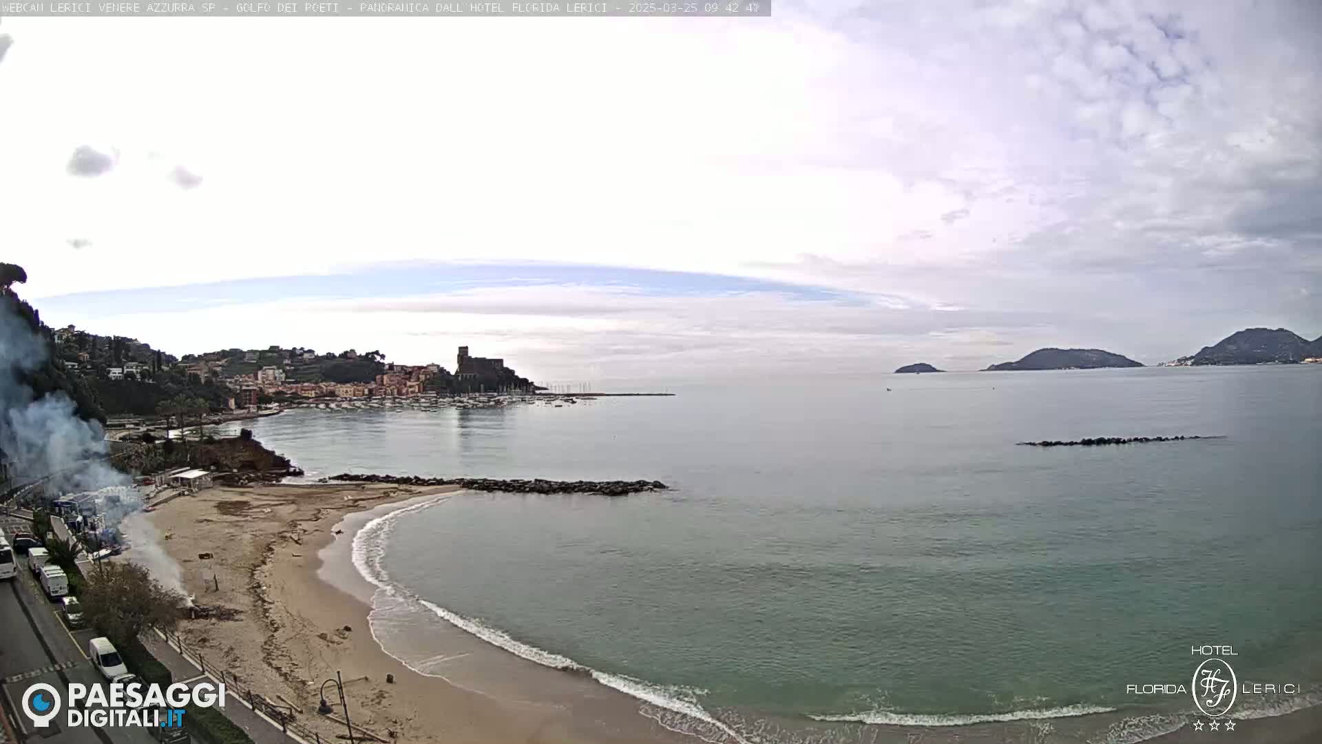 A sandy beach with small waves laps against a rocky breakwater; in the background, a town is nestled beside a calm bay, under a mostly cloudy sky.