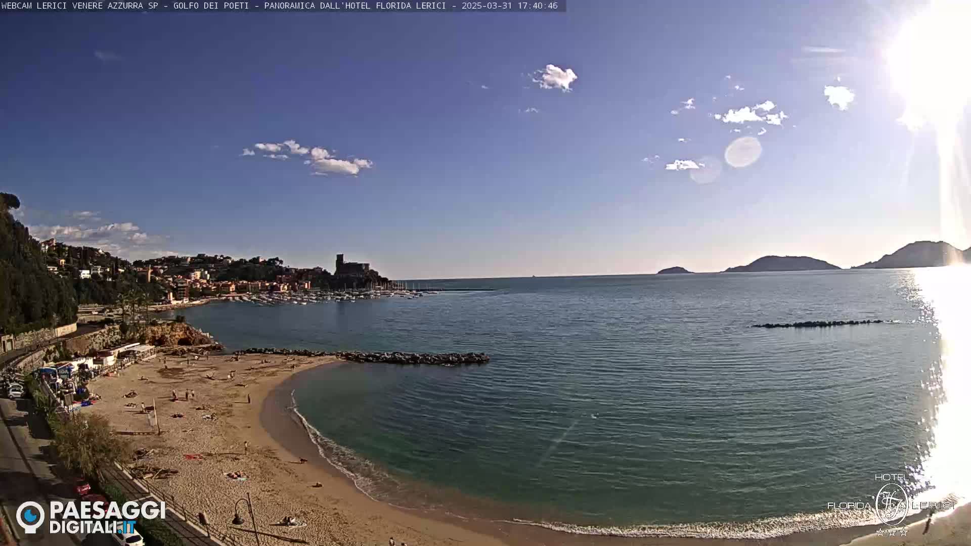 A sunny day reveals a secluded sandy beach curving into a calm bay, with a town and harbor nestled against a hillside in the background.