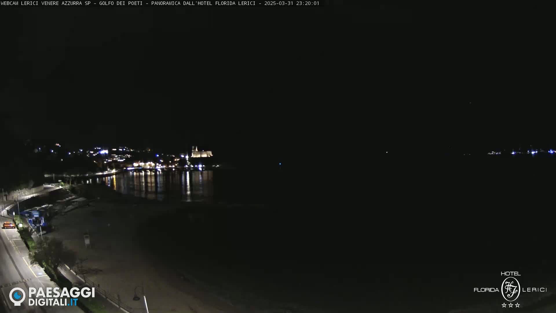 A nighttime view of a dark, calm bay with a town's lights reflecting on the water, a beach, and a road visible in the foreground.
