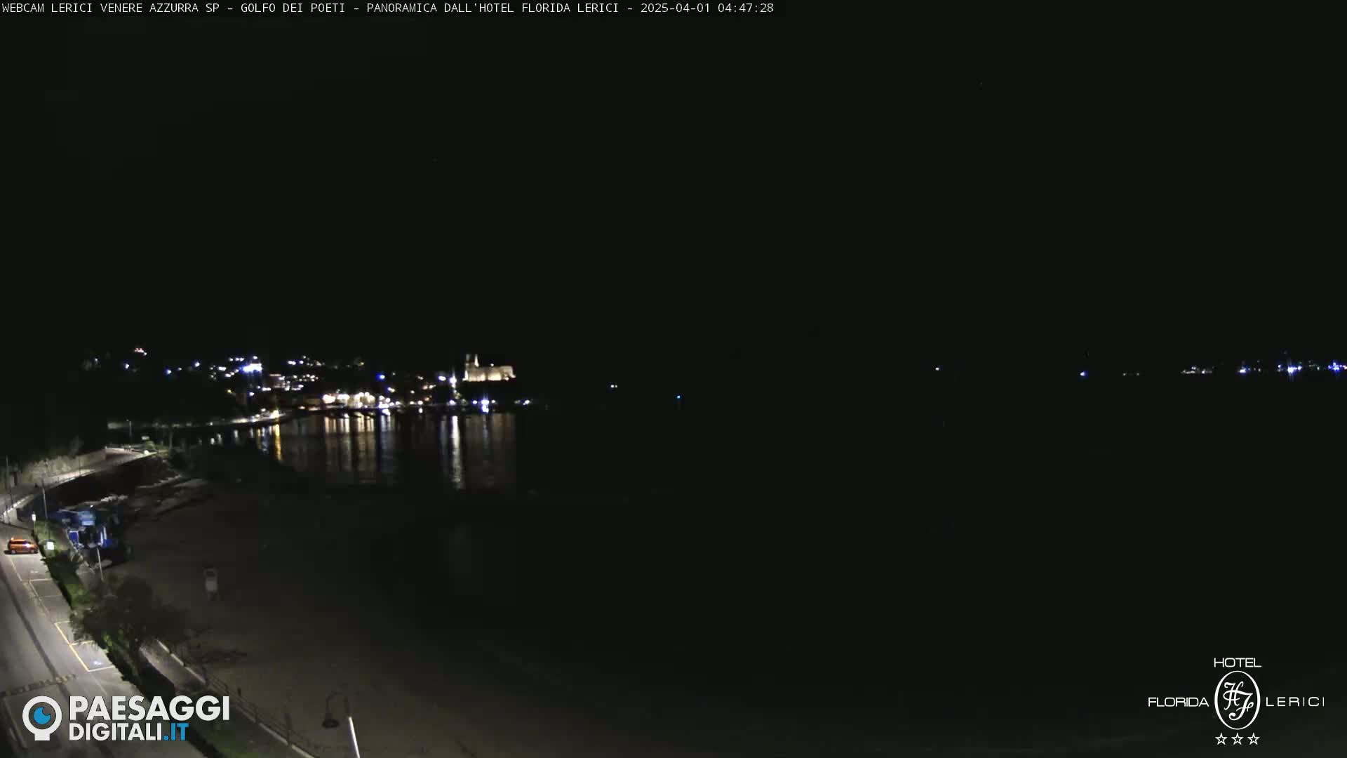 A nighttime view of a dark shoreline and calm water reflecting lights from a town across the bay.