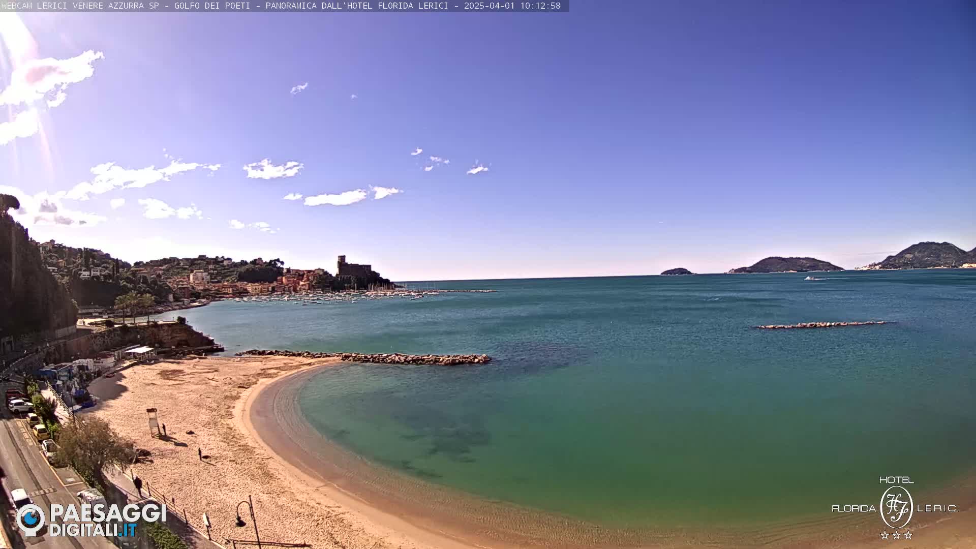 A sandy beach curves into a calm, teal-colored sea, with a town and distant hills under a mostly clear, sunny sky.
