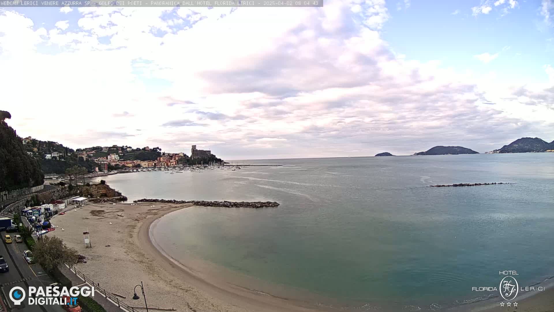 A sandy beach curves into a calm, teal-colored bay, with a town and castle visible across the water under a partly cloudy sky.