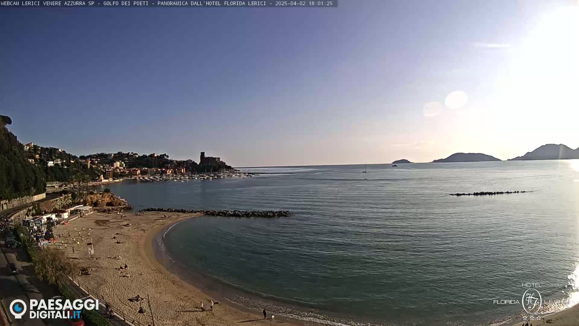 A sandy beach curves into a calm bay, with a town and distant hills visible across the water under a clear sky.