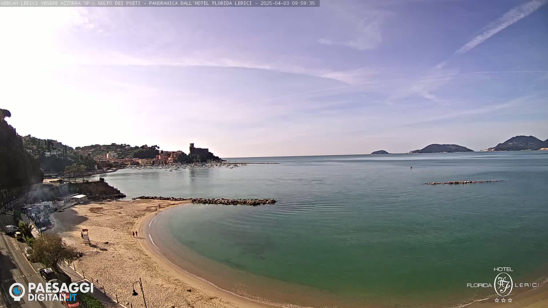 A sandy beach curves into a calm, teal sea, backed by a town nestled against green hills under a mostly clear sky.