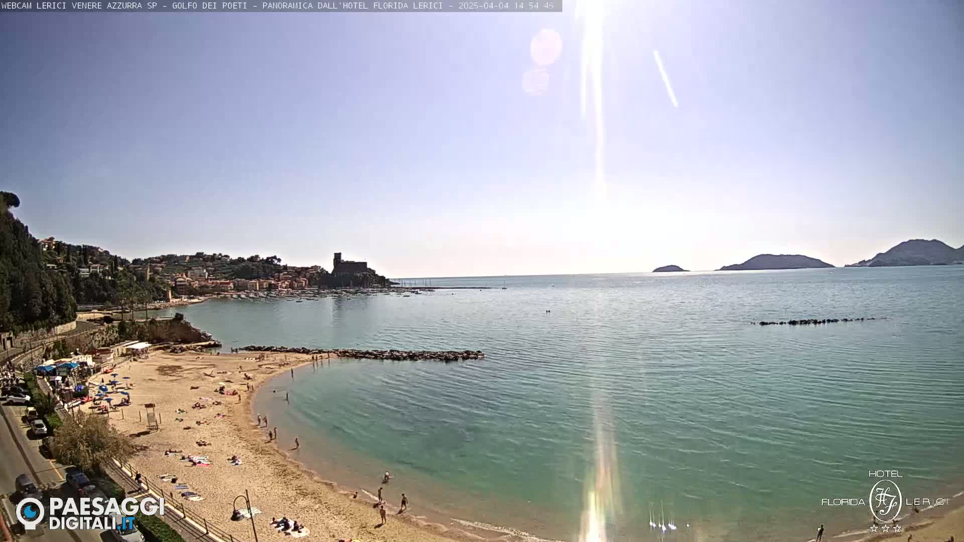 A sunny beach with people sunbathing and swimming, overlooks a calm bay with a town and distant hills in the background.