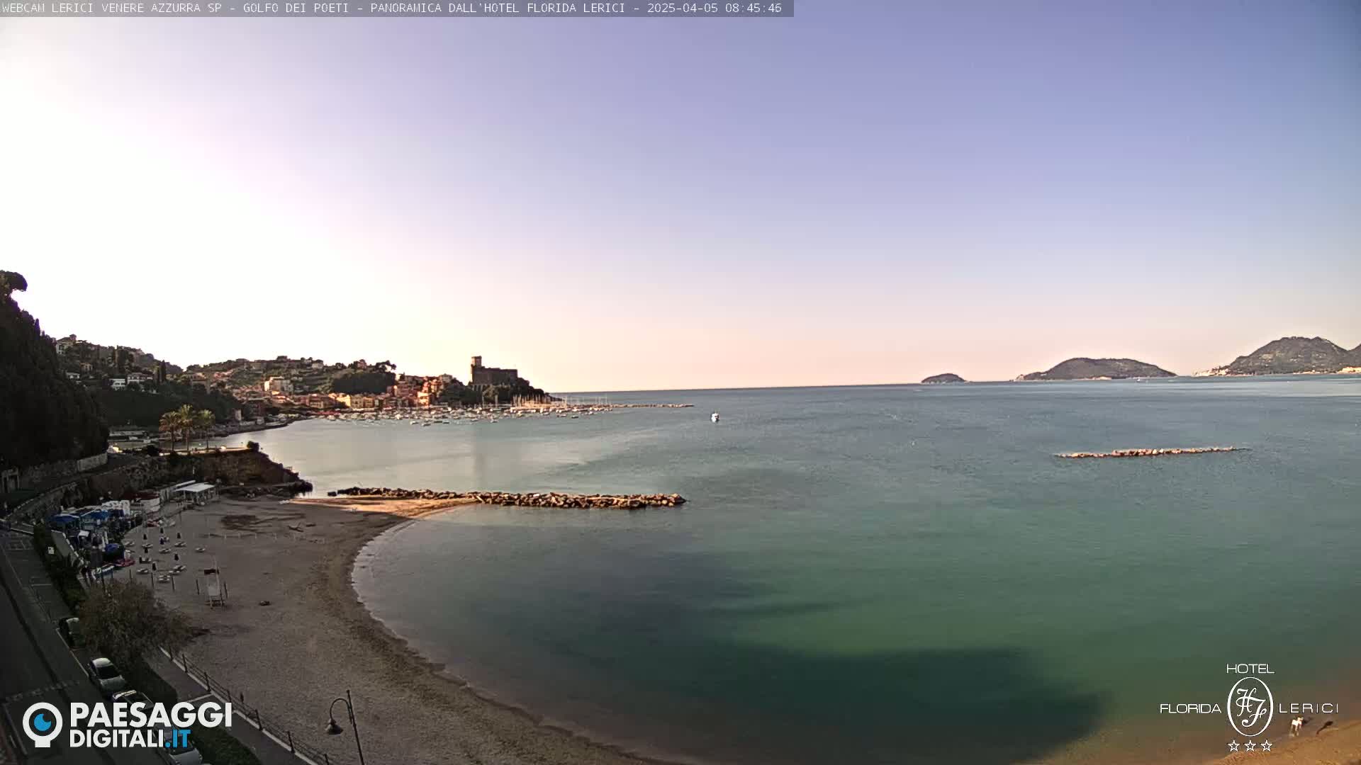 A tranquil beach scene with calm, teal-colored water, a sandy shoreline, and a distant town nestled against hills under a clear, light-blue sky.
