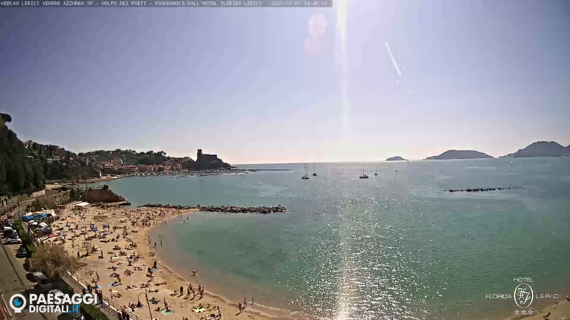 A sunny beach scene shows a crowded sandy beach next to calm, clear water with boats in the distance and hills in the background.